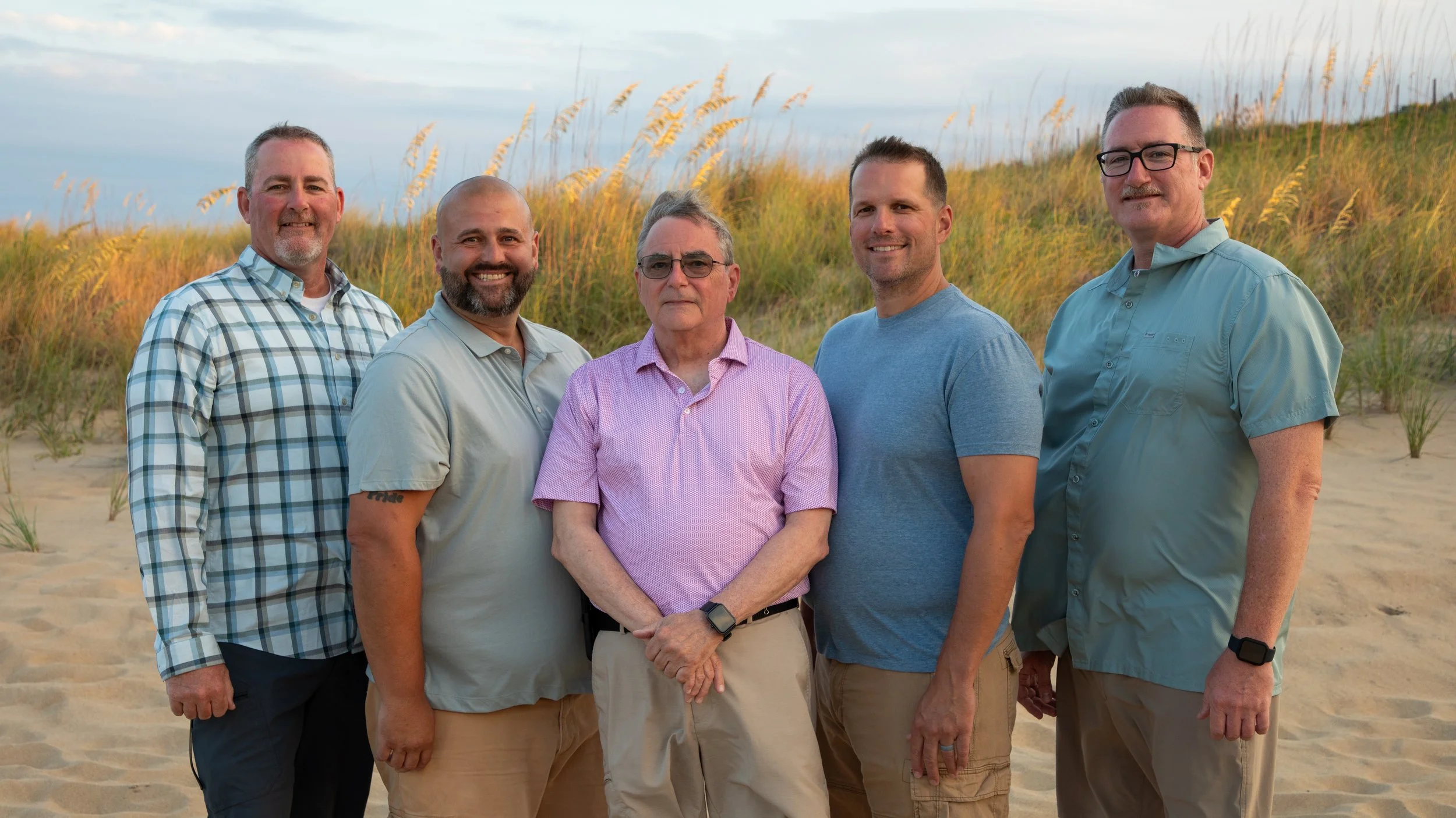 Photo & Video by Will Locke Family Photography in Virginia Beach, VA. Six men standing on a sandy beach with tall beach grass and a cloudy sky in the background.