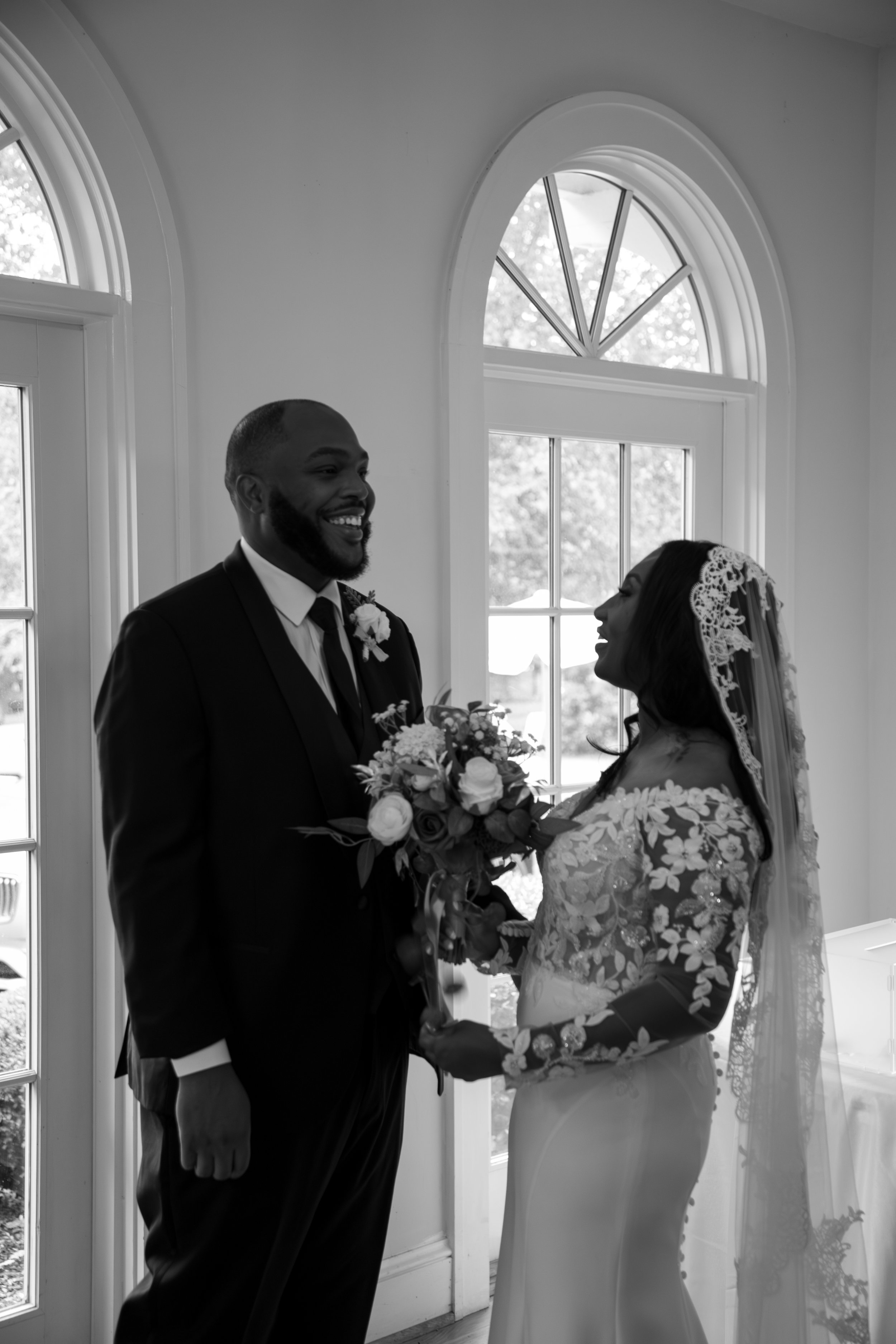 Wedding Photography by Will Locke. A bride and groom exchanging vows indoors, holding a bouquet, with large windows in the background.