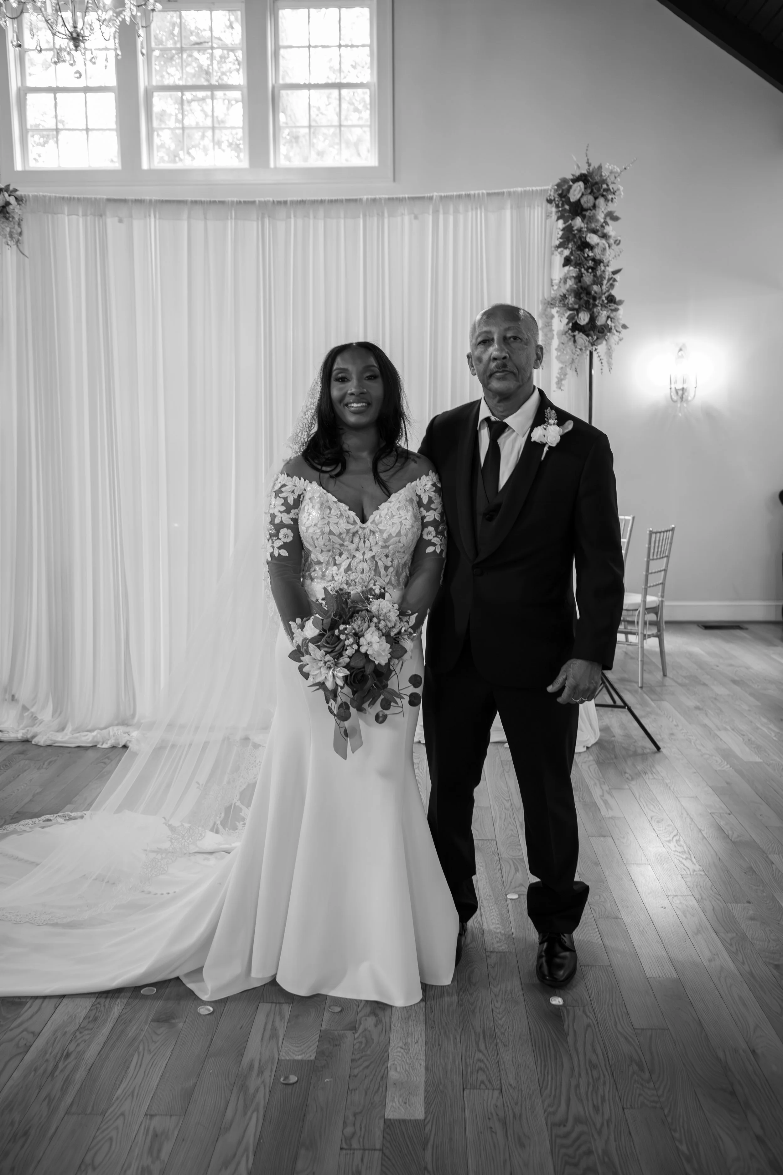 Photo & Video by Will Locke Wedding Photography at the Woman's Club of Portsmouth in Virginia. A bride in a lace wedding dress holding a bouquet stands next to a man in a suit inside a decorated venue with floral arrangements and a draped backdrop.