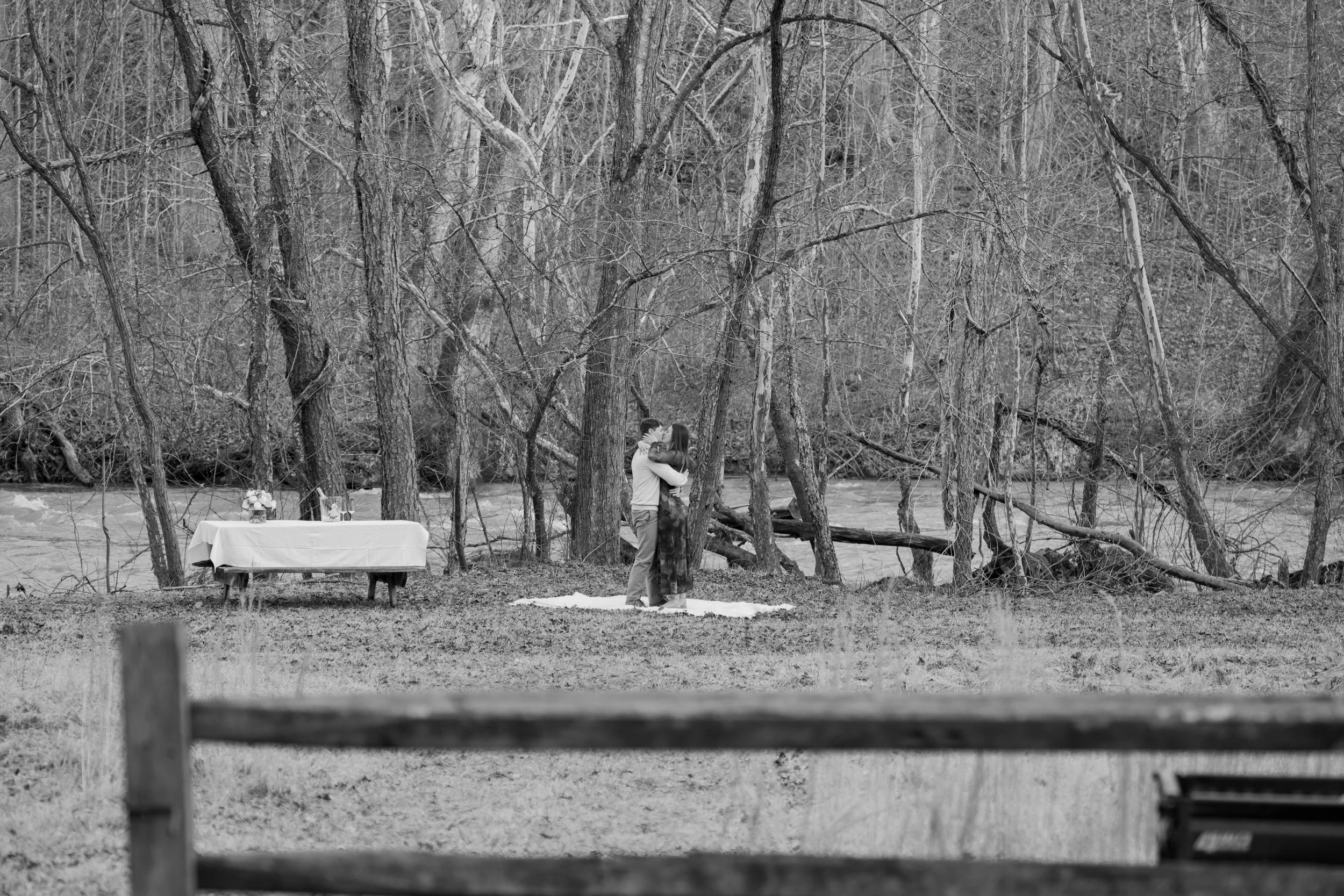 Engagement Photography by Will Locke near Richmond, VA in Montpelier. A couple hugging each other outdoors near a river, standing on a white blanket on the ground, surrounded by leafless trees in a forested area.