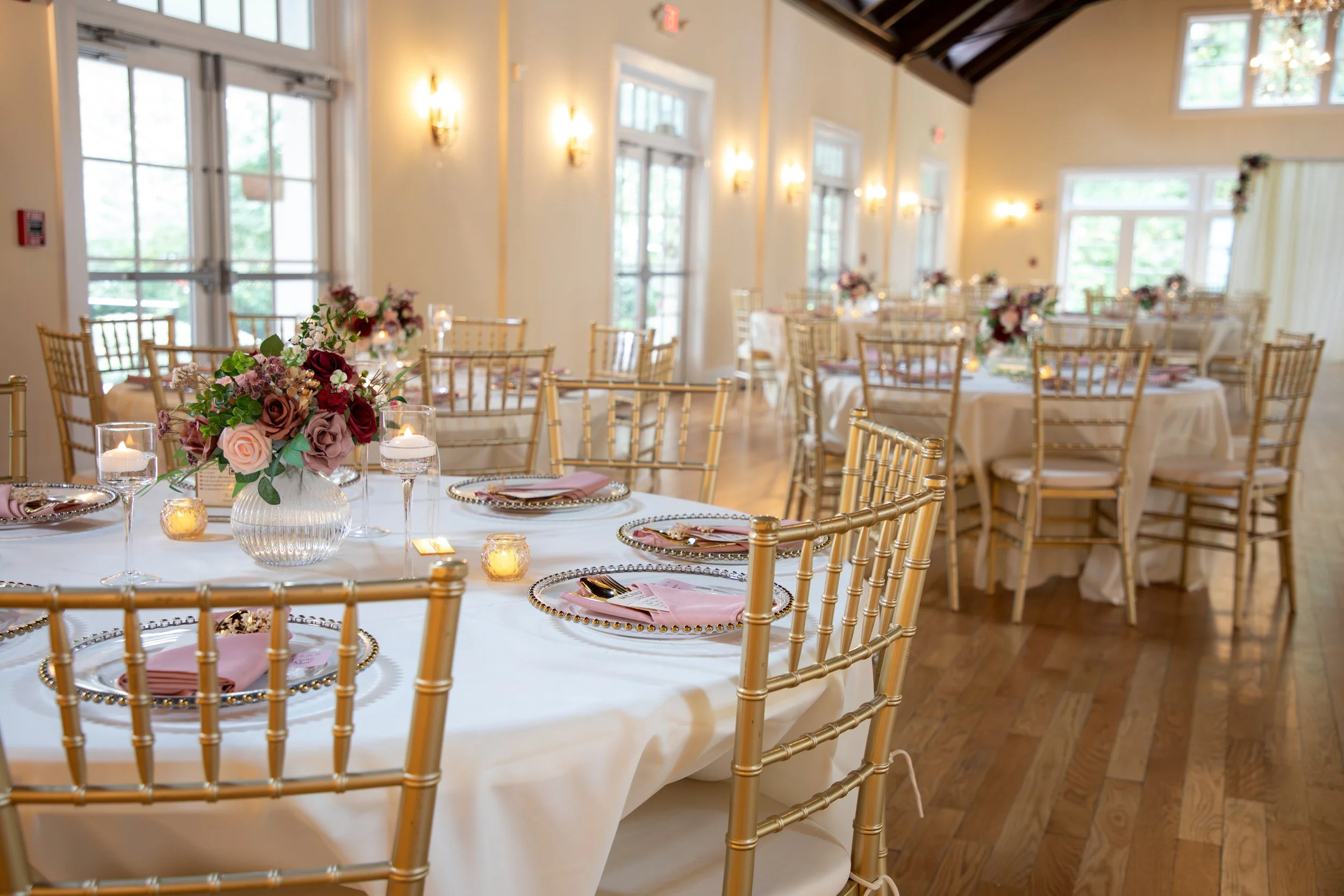 Wedding Photography by Will Locke. Elegant event space decorated with round tables covered in white tablecloths, gold chairs, floral centerpieces, candles, and place settings, with large windows providing natural light.