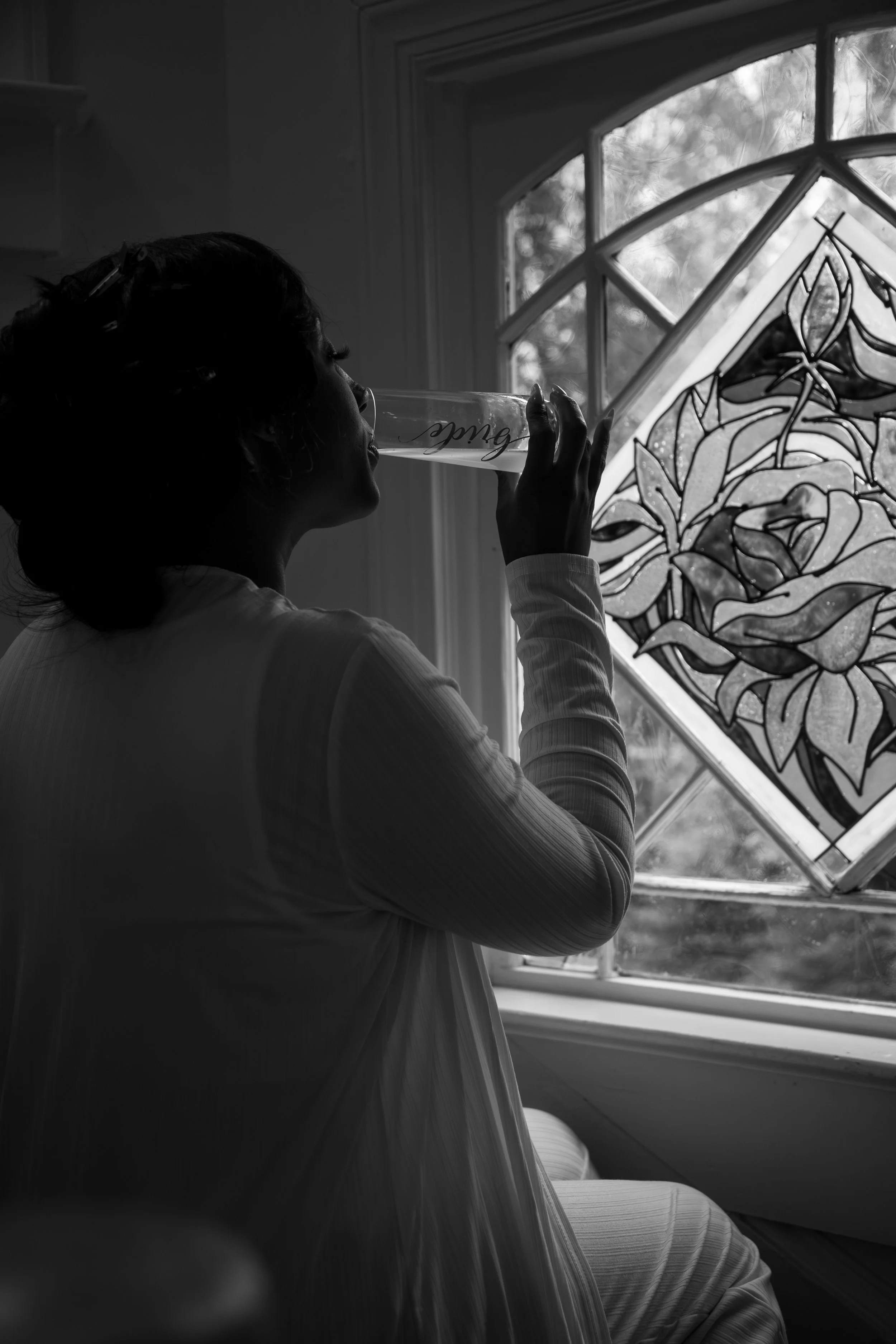 Wedding Photography by Will Locke. A woman in a long-sleeved top drinks from a glass with script writing, sitting near a stained glass window with floral design, in a dimly lit room.
