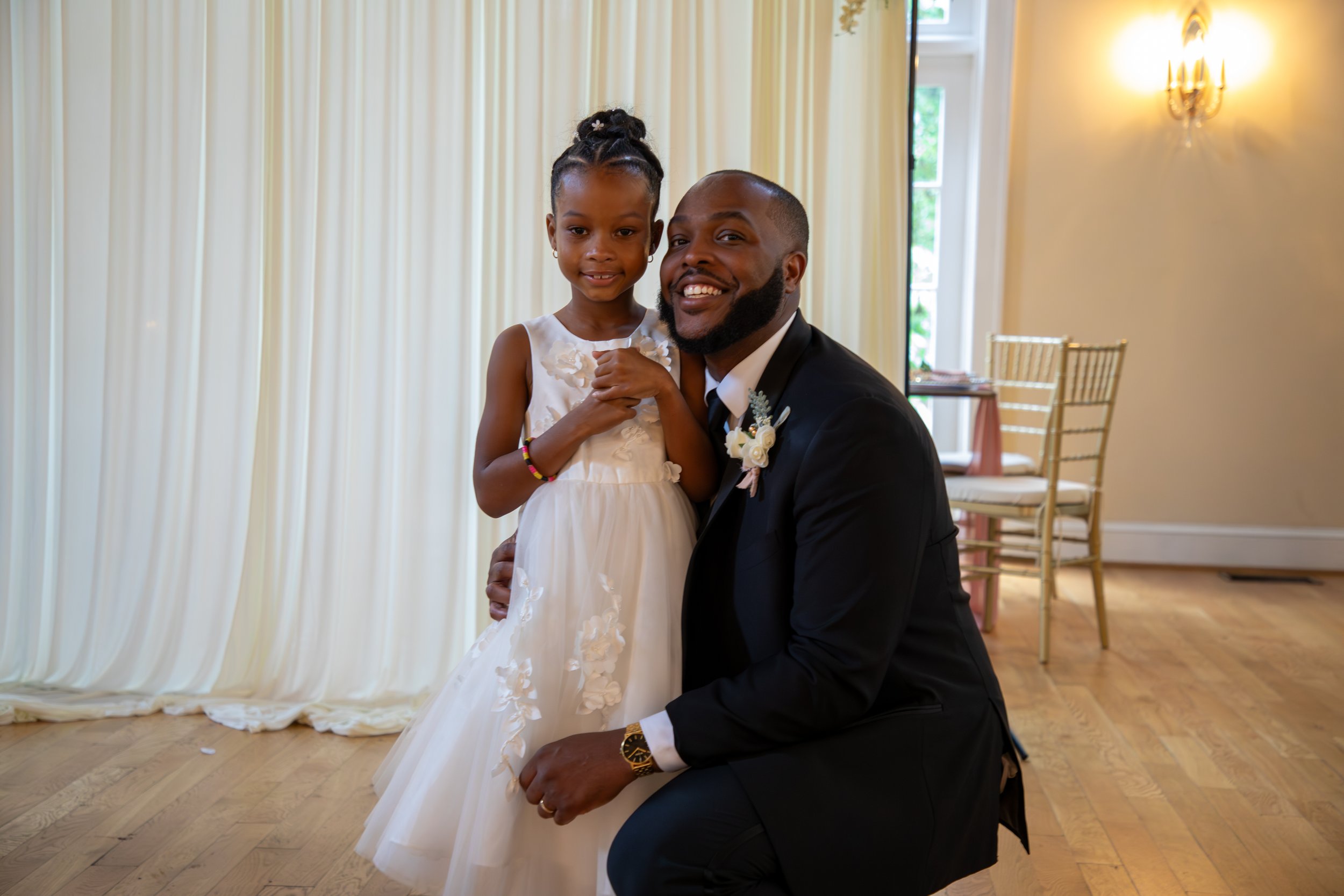 Photo & Video by Will Locke Wedding Photography at the Woman's Club of Portsmouth in Virginia. A man in a black suit and a young girl in a white dress are smiling and posing together in a decorated indoor setting, possibly at a wedding or celebration