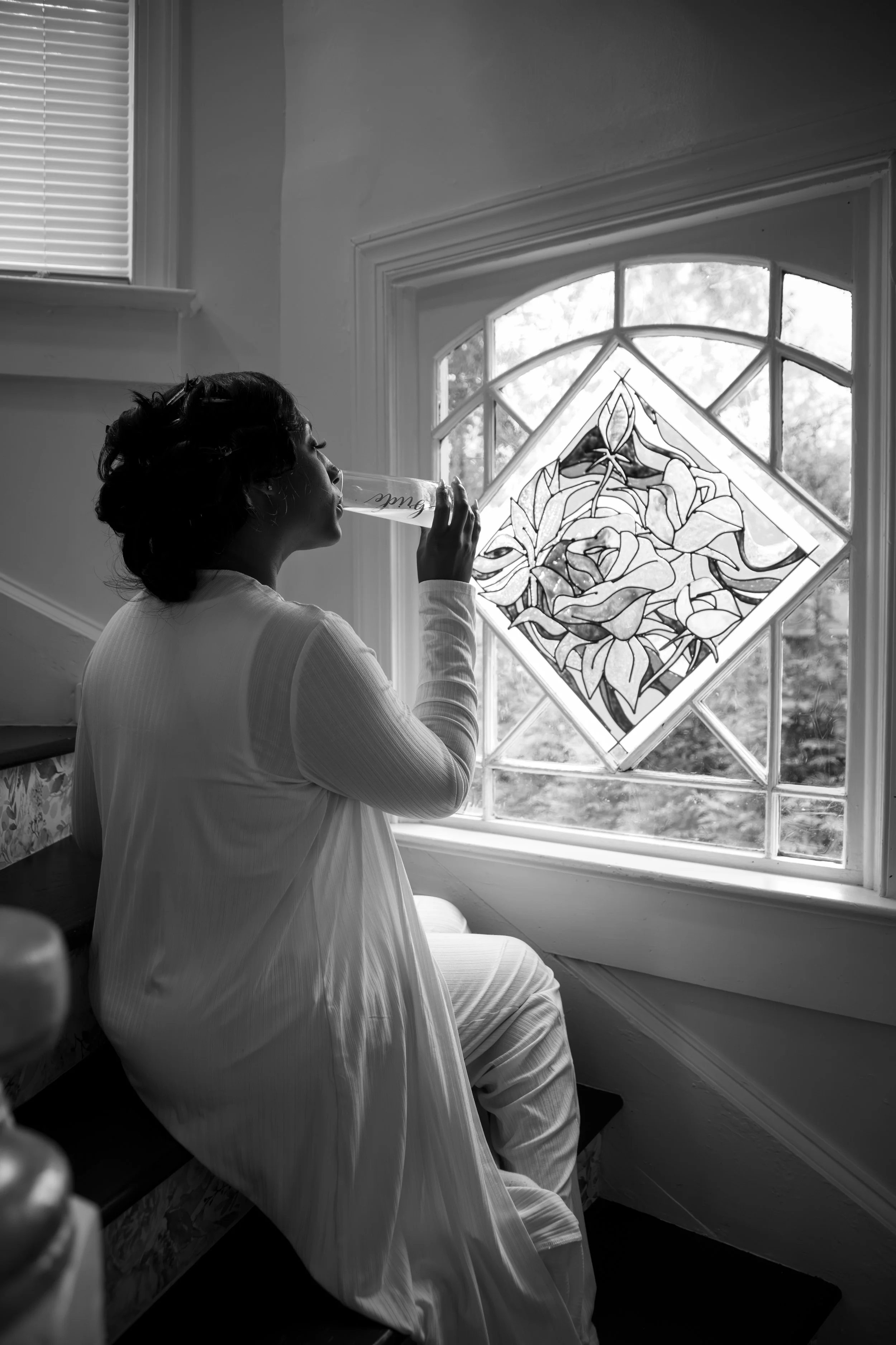 Photo & Video by Will Locke Wedding Photography at the Bridgerton House in Portsmouth, VA. A woman in a long dress sitting on stairs next to a stained glass window with floral design, blowing into a glass tube.