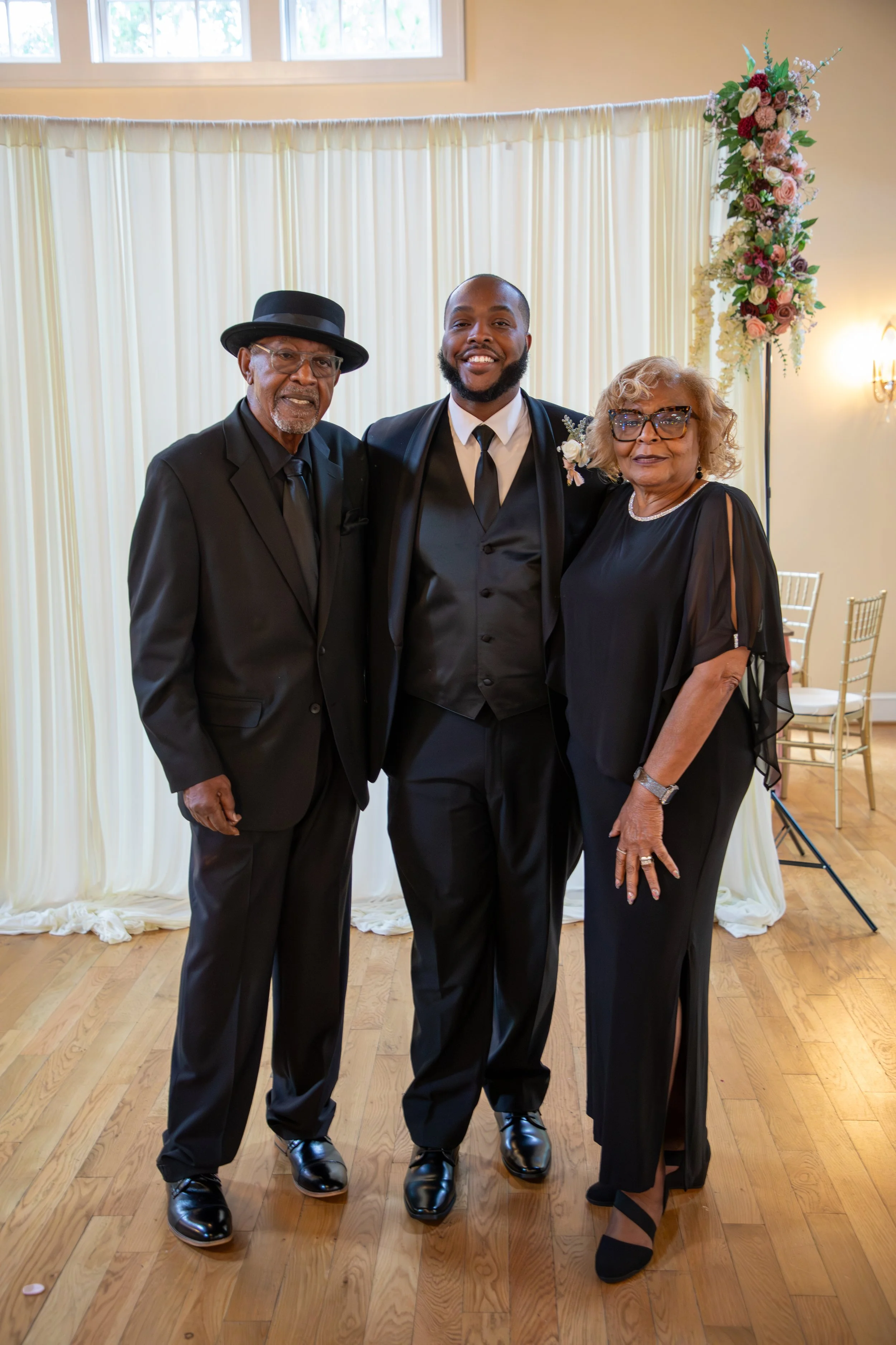 Photo & Video by Will Locke Wedding Photography at the Woman's Club of Portsmouth in VA. Three people in formal attire at a wedding, standing indoors with a cream-colored backdrop and a floral arrangement in the corner.
