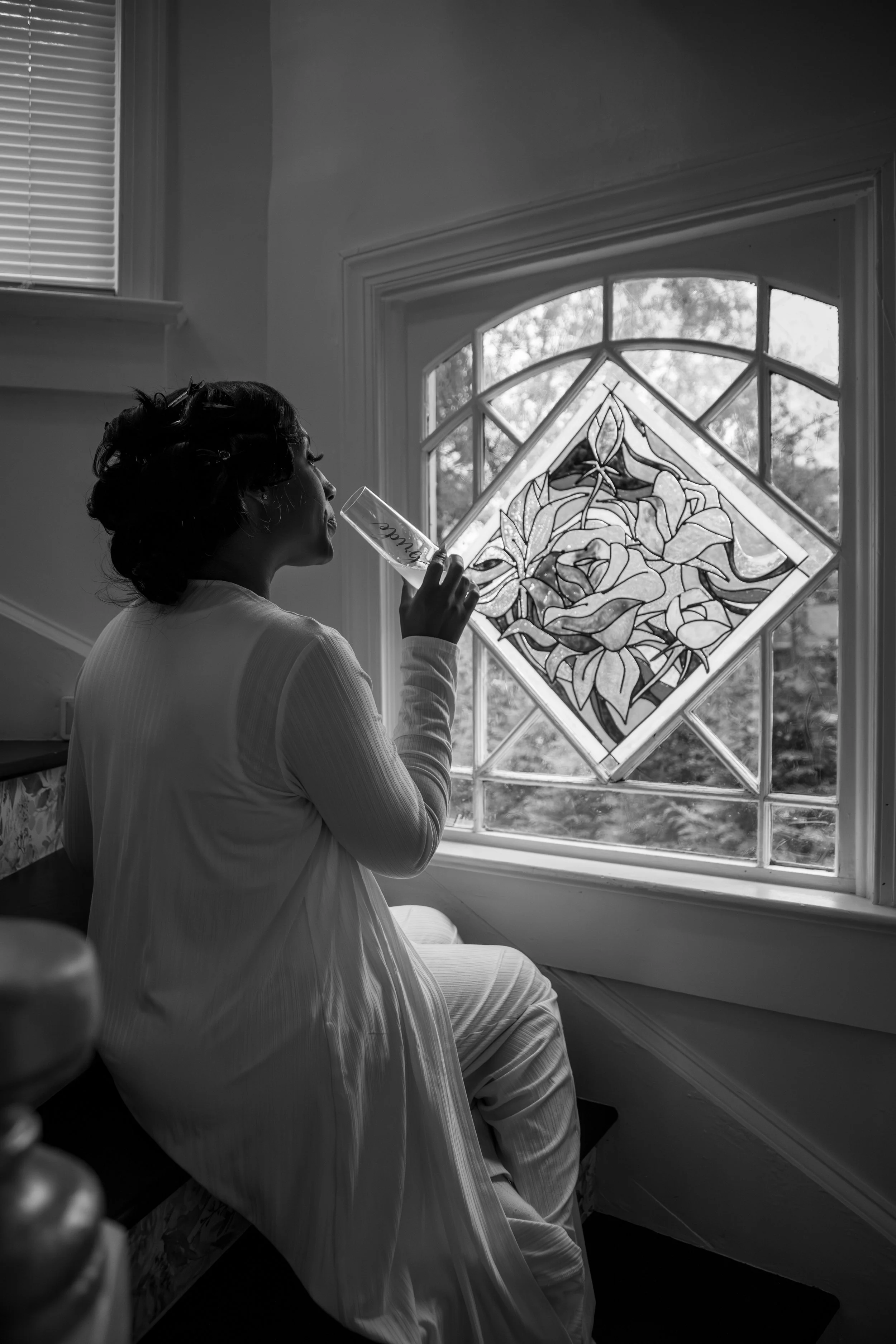 Photo & Video by Will Locke Wedding Photography at the Bridgerton House in Portsmouth, VA. A woman with short dark hair, sitting on a bench inside a house, drinking from a glass, near a large window with stained glass art, in black and white.