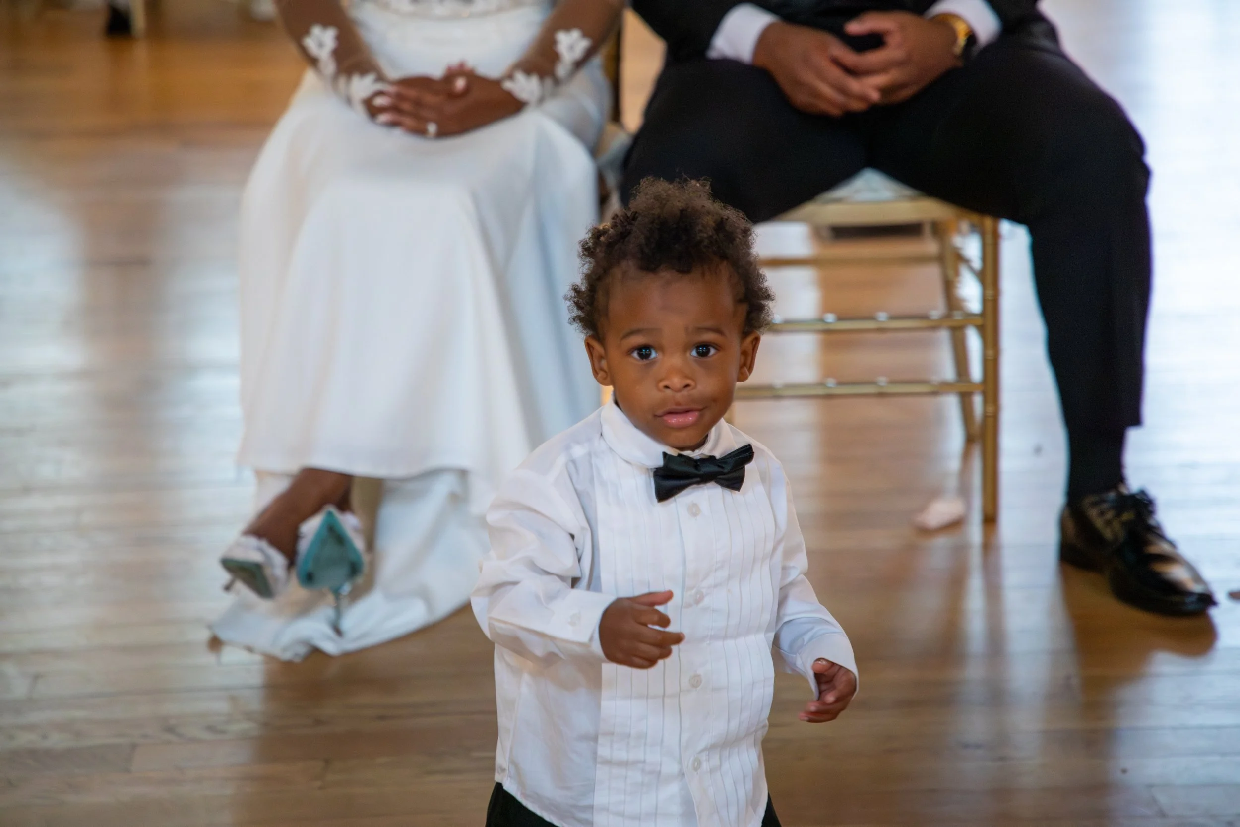 Photo & Video by Will Locke Wedding Photography at the Woman's Club of Portsmouth. A young boy with curly hair wearing a white dress shirt with a black bow tie, standing in a room with a wooden floor, looking at the camera with a curious expression.