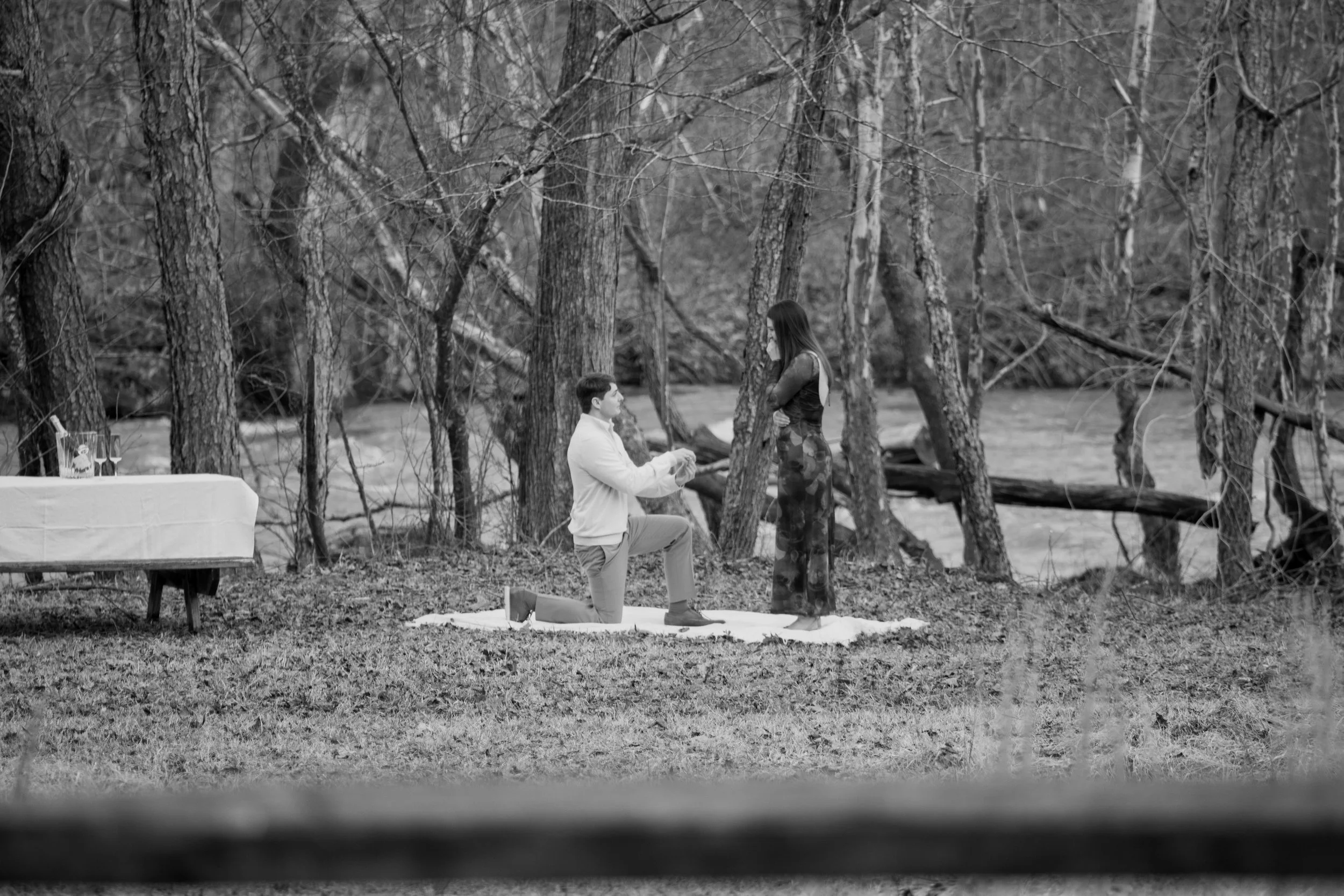 Engagement Photography by Will Locke near Richmond, VA in Montpelier. A man proposing to a woman outdoors in a wooded area near a river, with a small table set with drinks nearby.