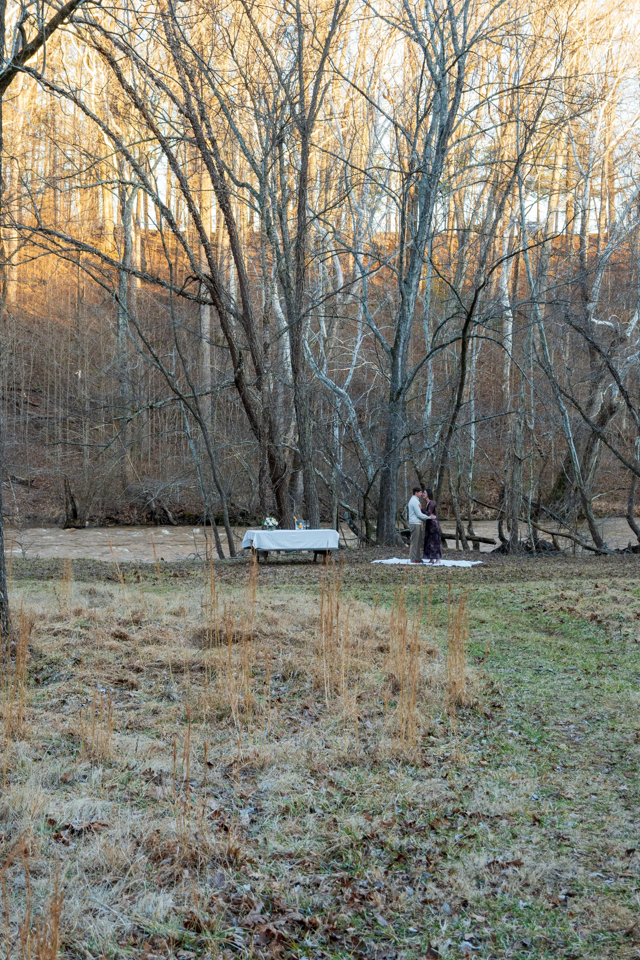 Engagement Photography by Will Locke near Richmond, VA in Montpelier. A couple hugging each other outdoors near a river, standing on a white blanket on the ground, surrounded by leafless trees in a forested area.