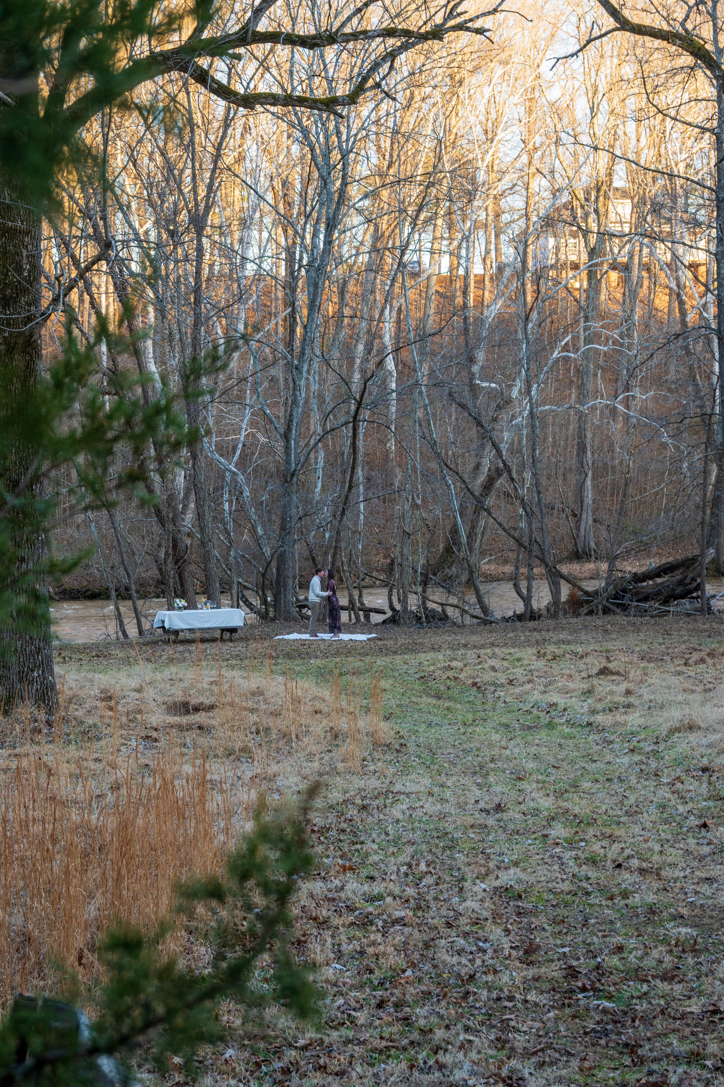 Engagement Photography by Will Locke near Richmond, VA in Montpelier. A couple hugging each other outdoors near a river, standing on a white blanket on the ground, surrounded by leafless trees in a forested area.