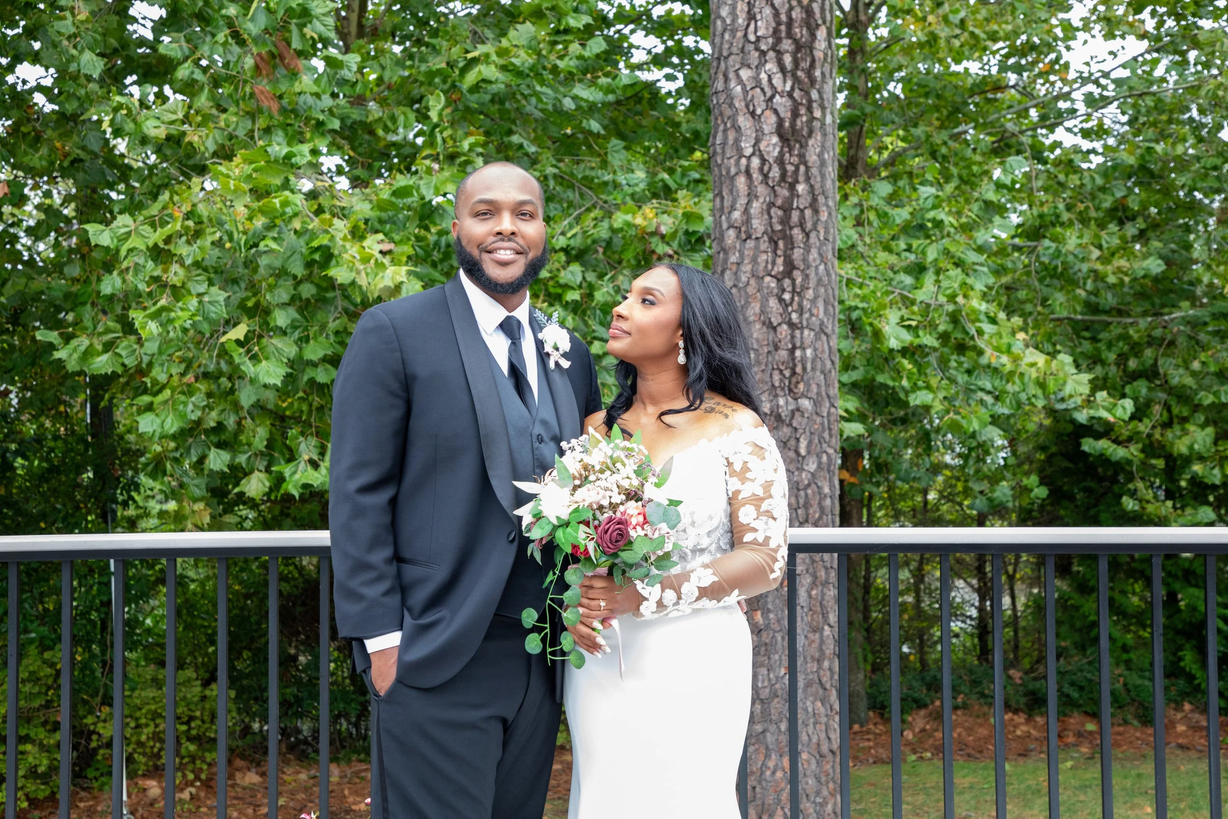Photo & Video by Will Locke Wedding Photography at the Woman's Club of Portsmouth. A newlywed couple posing outdoors with a lush green background, the groom in a black tuxedo and the bride in a white lace wedding dress holding a bouquet of flowers.