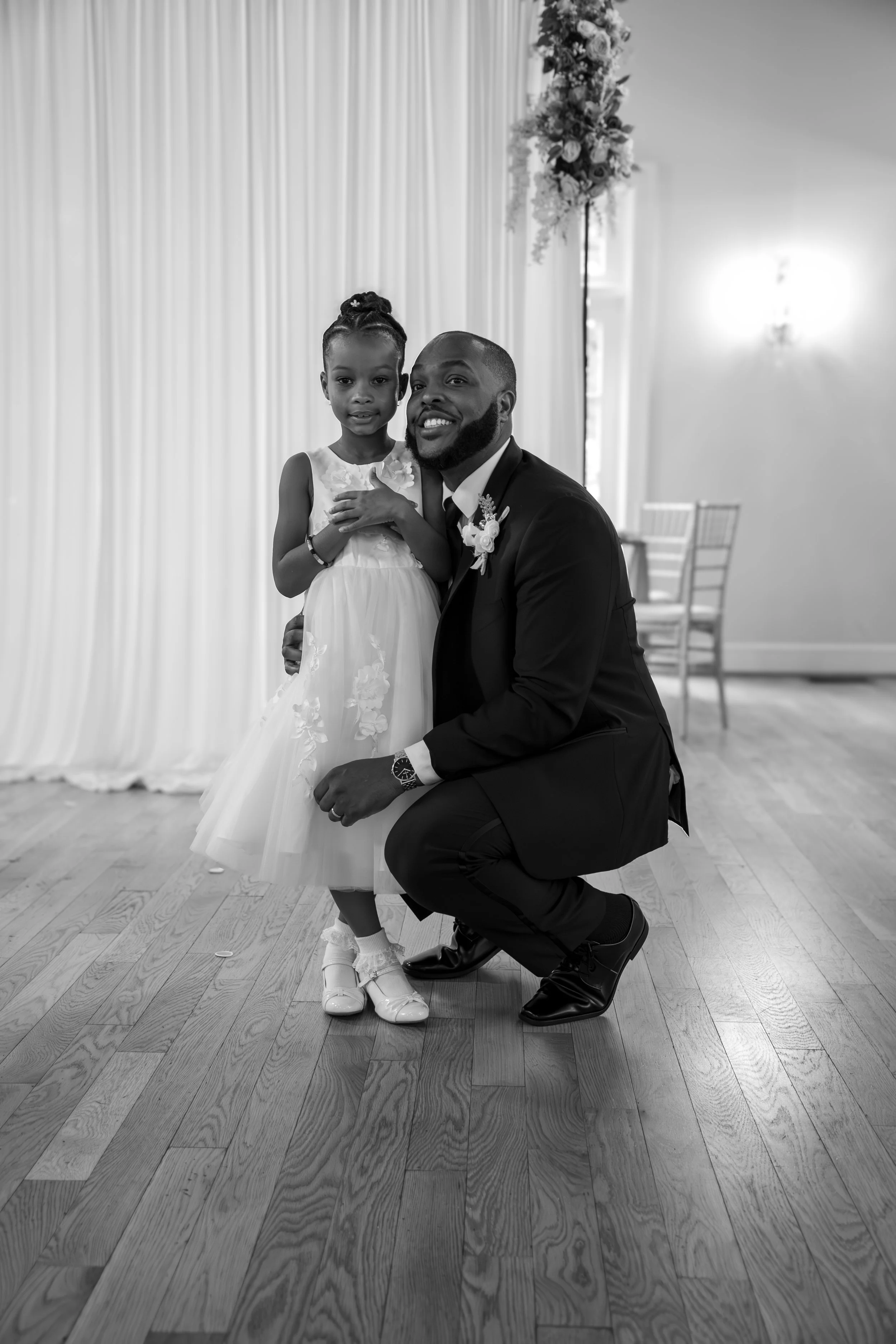 Photo & Video by Will Locke Wedding Photography at the Woman's Club of Portsmouth in Virginia. A man and a young girl, dressed in formal attire, posing happily at a wedding reception with a decorated backdrop and wooden floor.