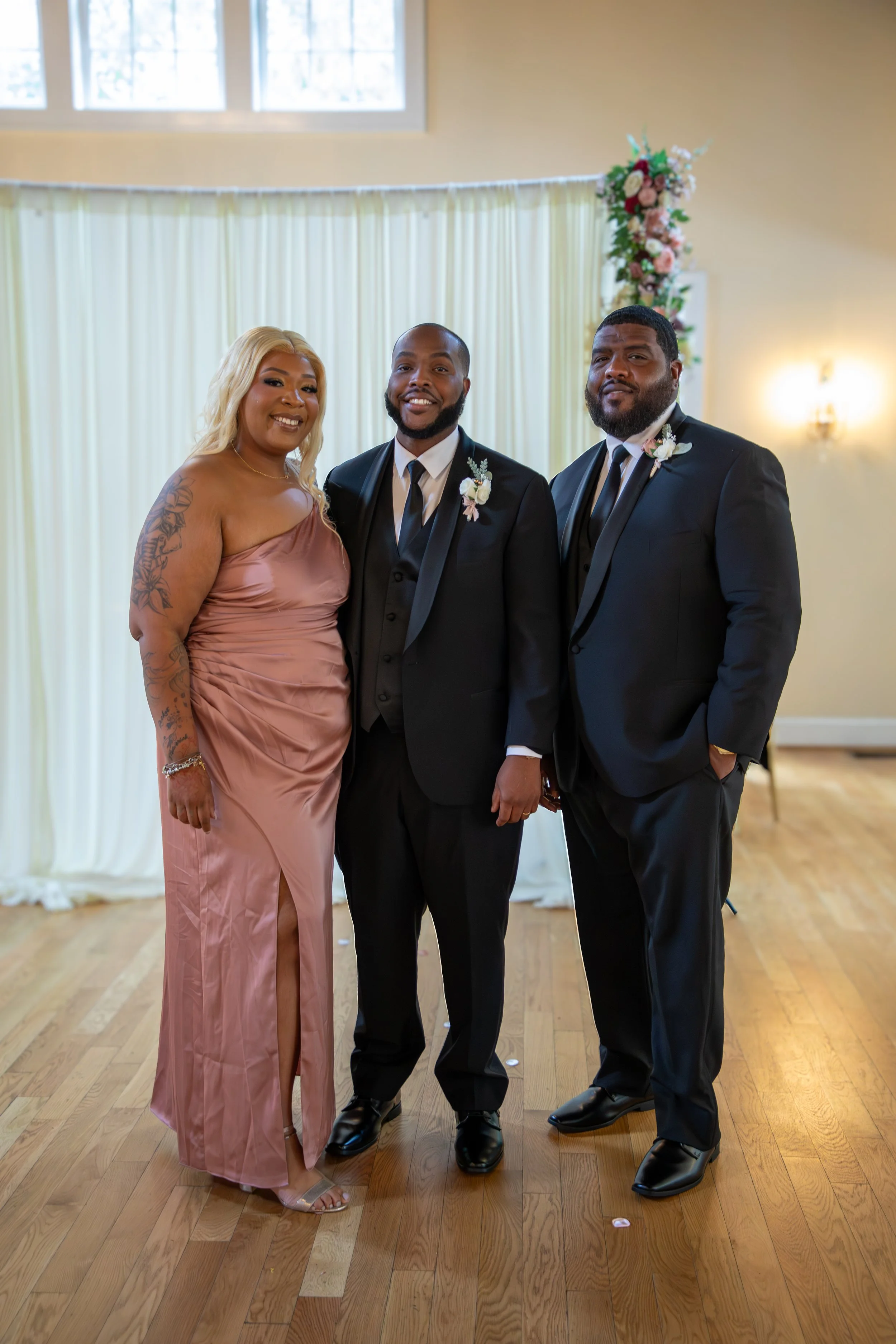 Photo & Video by Will Locke Wedding Photography at the Woman's Club of Portsmouth in Virginia. Three people standing together indoors at a formal event, possibly a wedding, with a cream-colored curtain and floral decoration behind them.