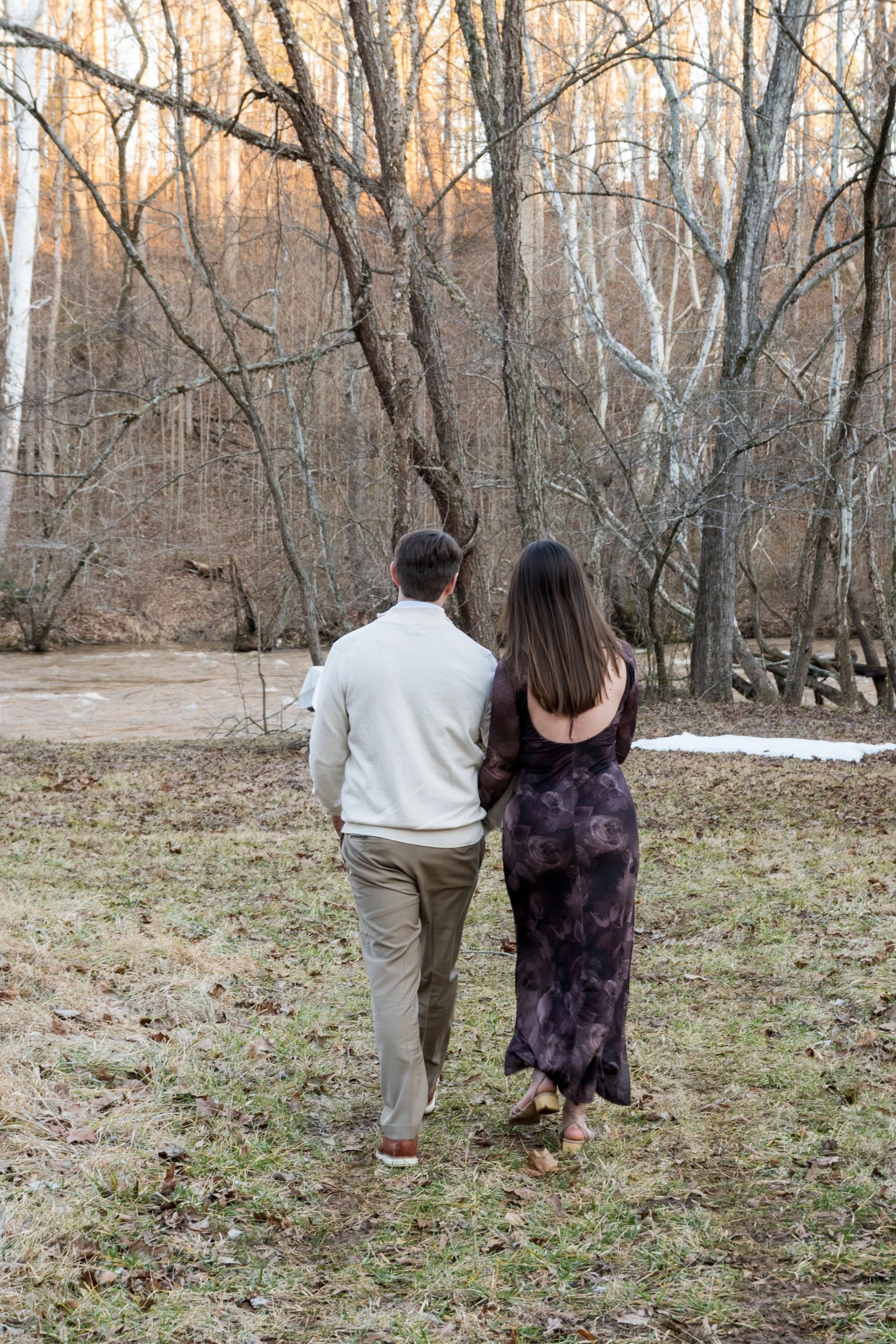 Engagement Photography by Will Locke near Richmond, VA in Montpelier. A couple walks to a white cloth on the ground near a wooded area with a stream on a fall day, with a table on the left side with white tablecloth.