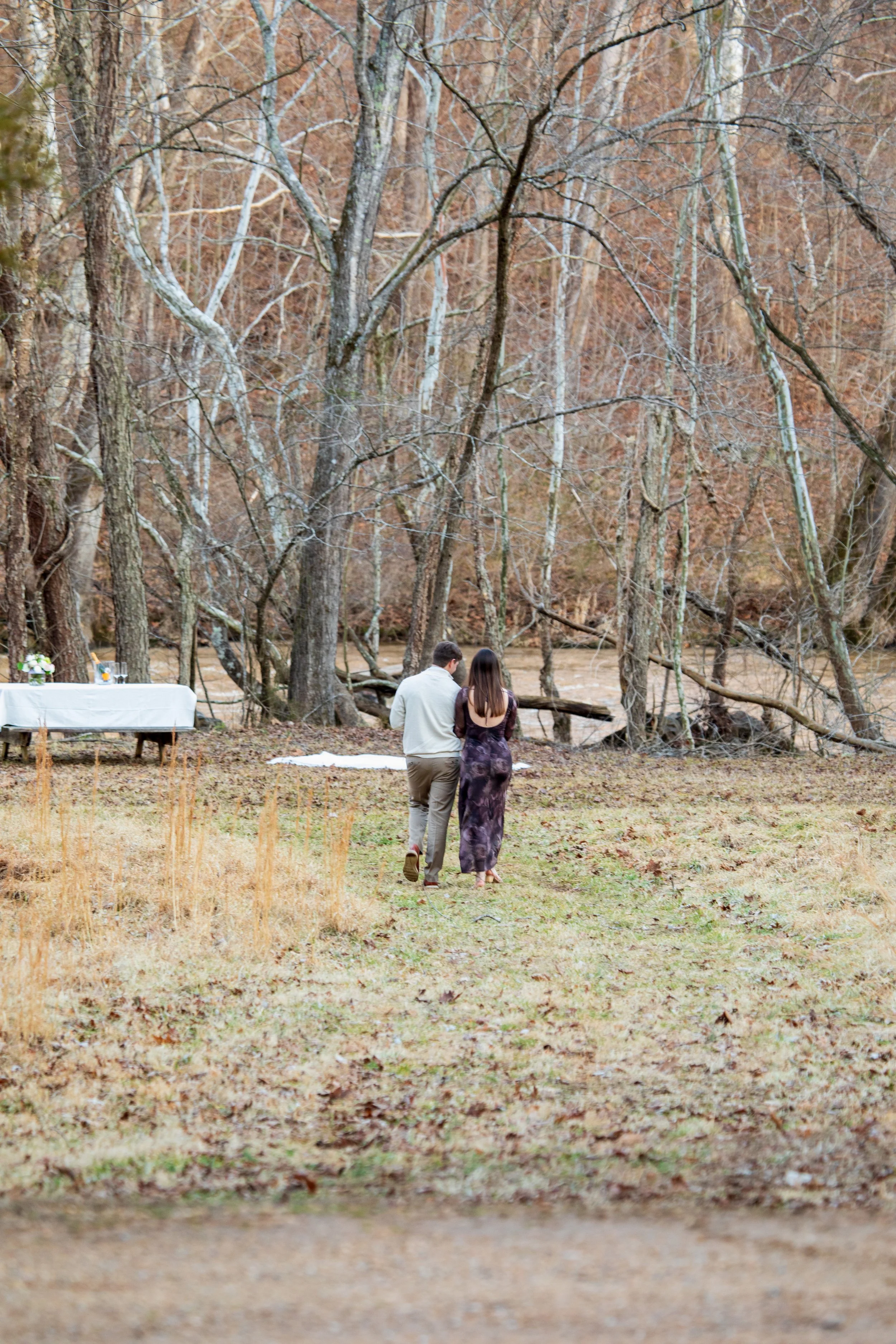 Engagement Photography by Will Locke near Richmond, VA in Montpelier. A couple walks together in a wooded outdoor area with leafless trees and a river in the background, near a picnic table covered with a white tablecloth.
