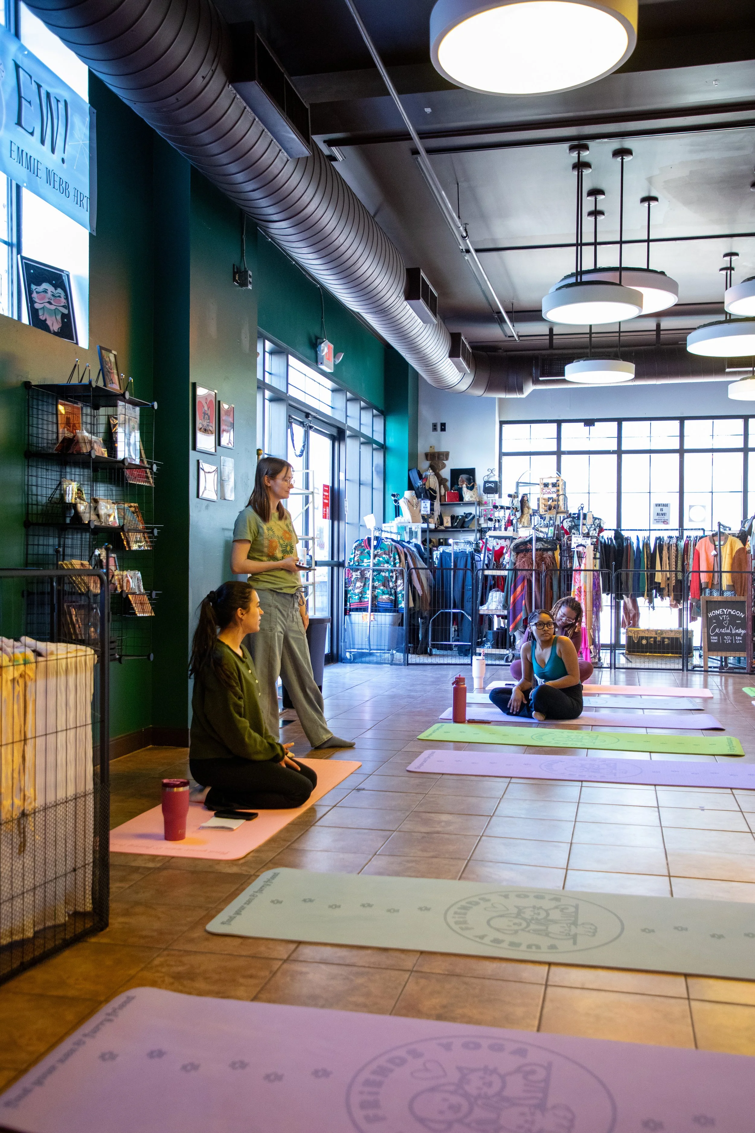 Marketing and Event Photography by Will Locke. Four women participating in a yoga class inside a retail store, with yoga mats laid out on the tiled floor, clothing displays in the background, and large windows allowing natural light.