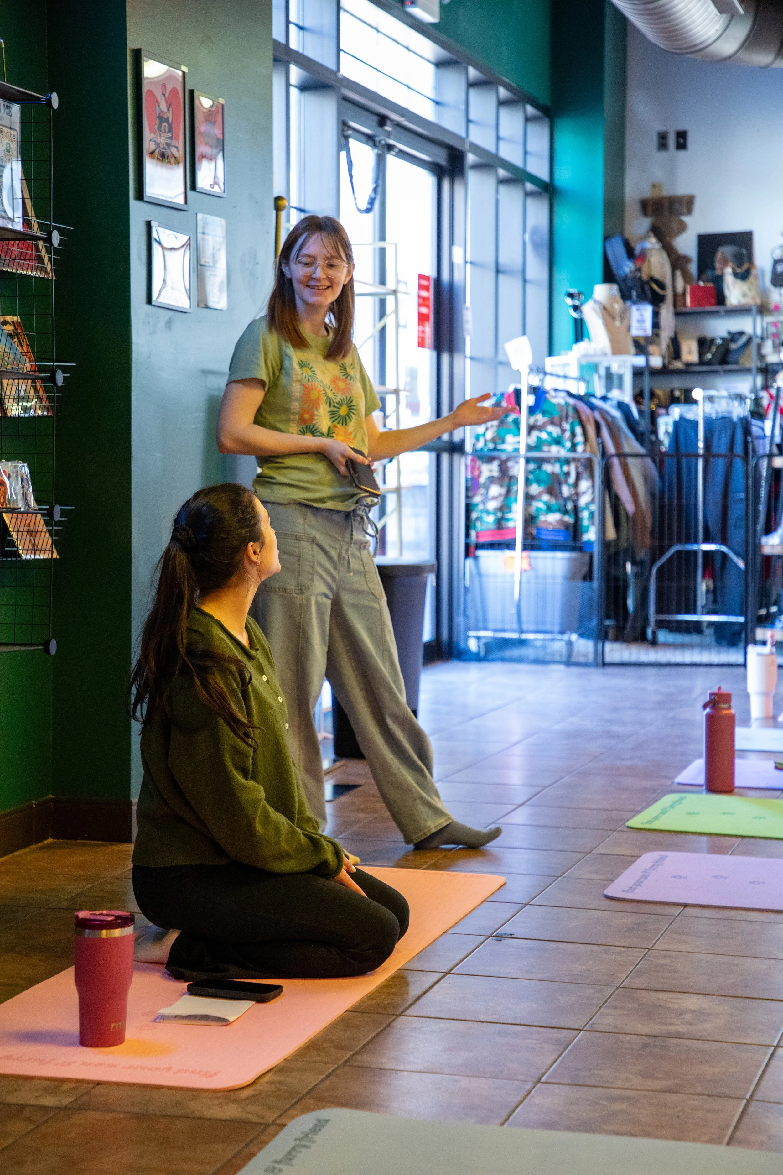 Marketing and Event Photography by Will Locke. Two women participating in a yoga class inside a retail store, with one standing and the other sitting on a yoga mat, surrounded by retail merchandise and sporting a casual outfit.