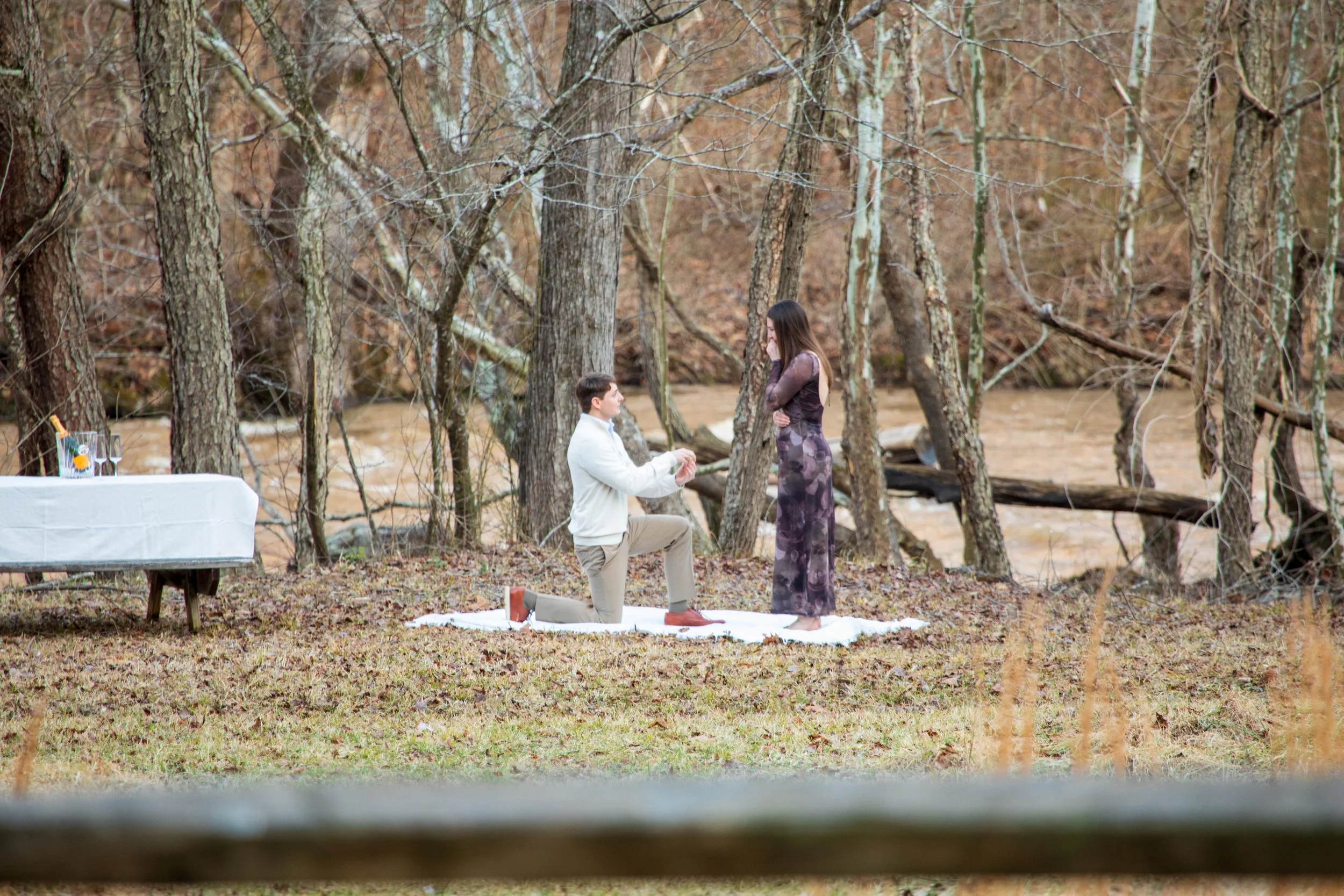 Engagement Photography by Will Locke near Richmond, VA in Montpelier. A man proposing to a woman outdoors in a wooded area near a river, with a small table set with drinks nearby.