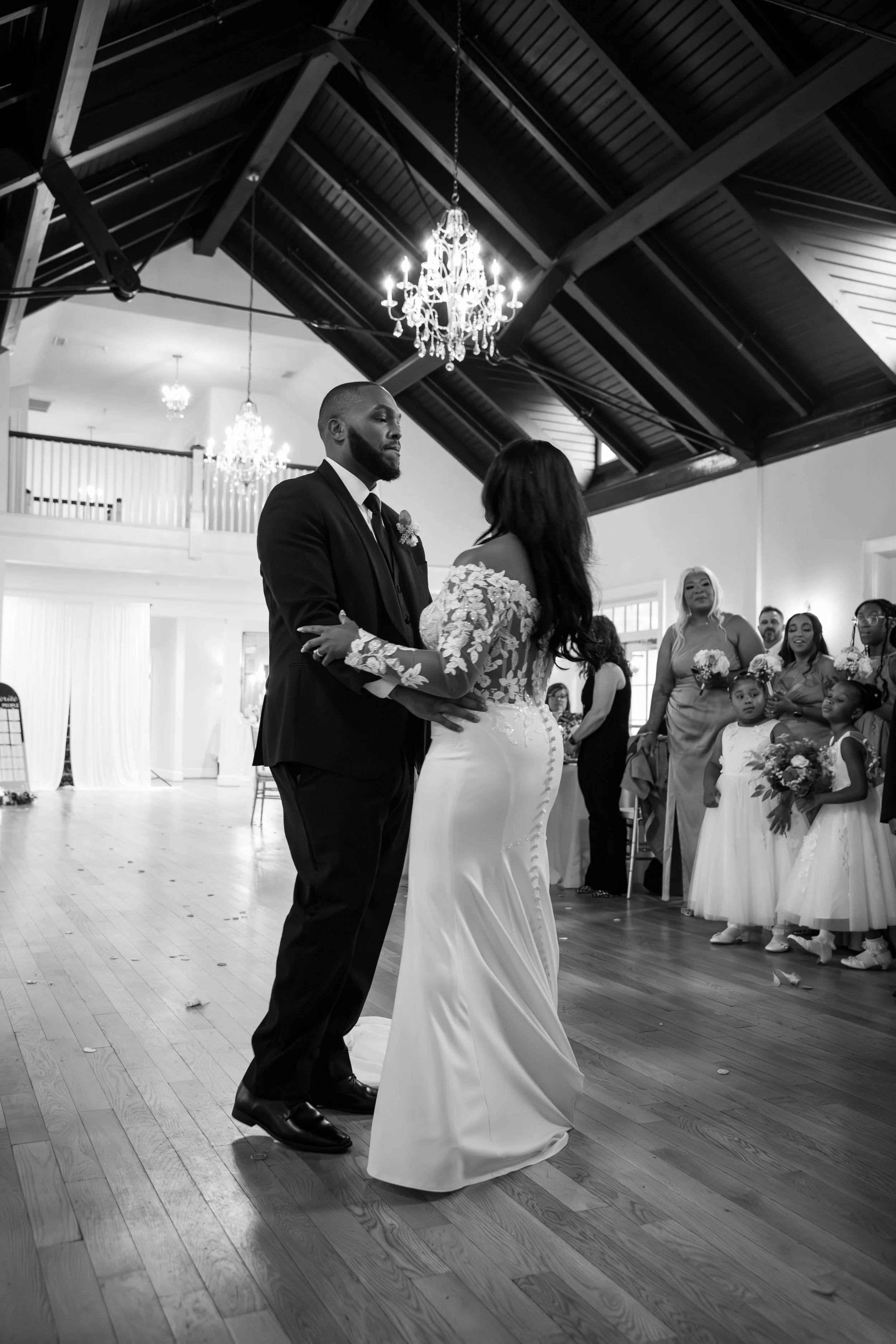 Wedding Photography by Will Locke. A black and white photo of a wedding couple dancing, surrounded by guests and flower girls, with chandeliers hanging from the ceiling.