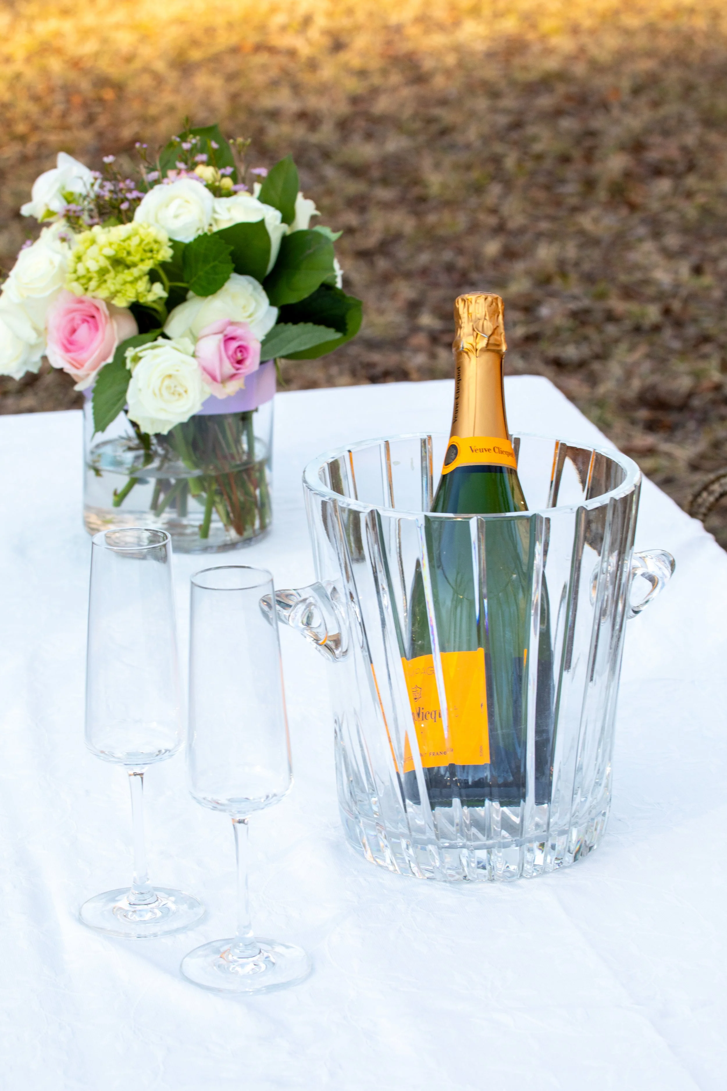 Engagement Photography by Will Locke near Richmond, VA in Montpelier. A bottle of Veuve Clicquot champagne in an ice bucket with two empty champagne flutes on a white table outdoors, with a flower arrangement. 