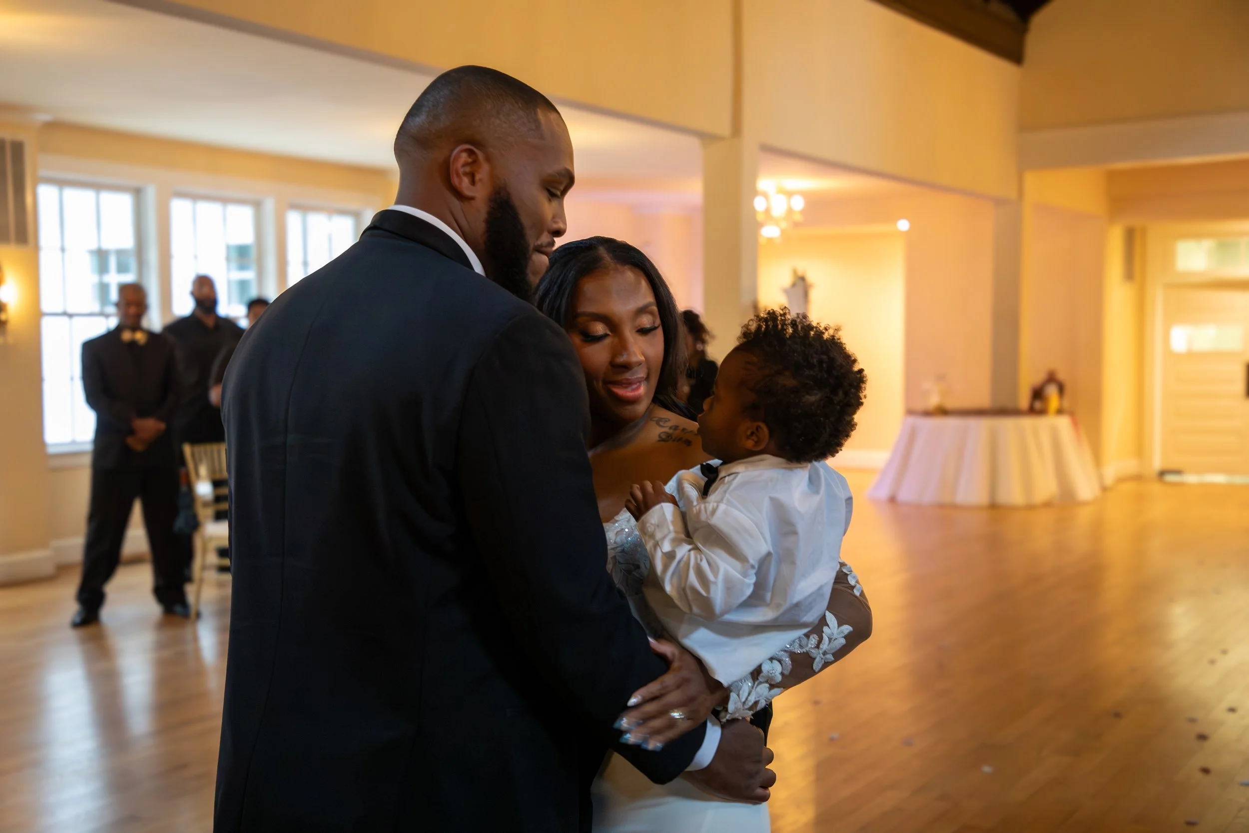 Wedding Photography by Will Locke. A wedding scene with a groom, bride, and a young child sharing a special moment inside a decorated venue with warm lighting.