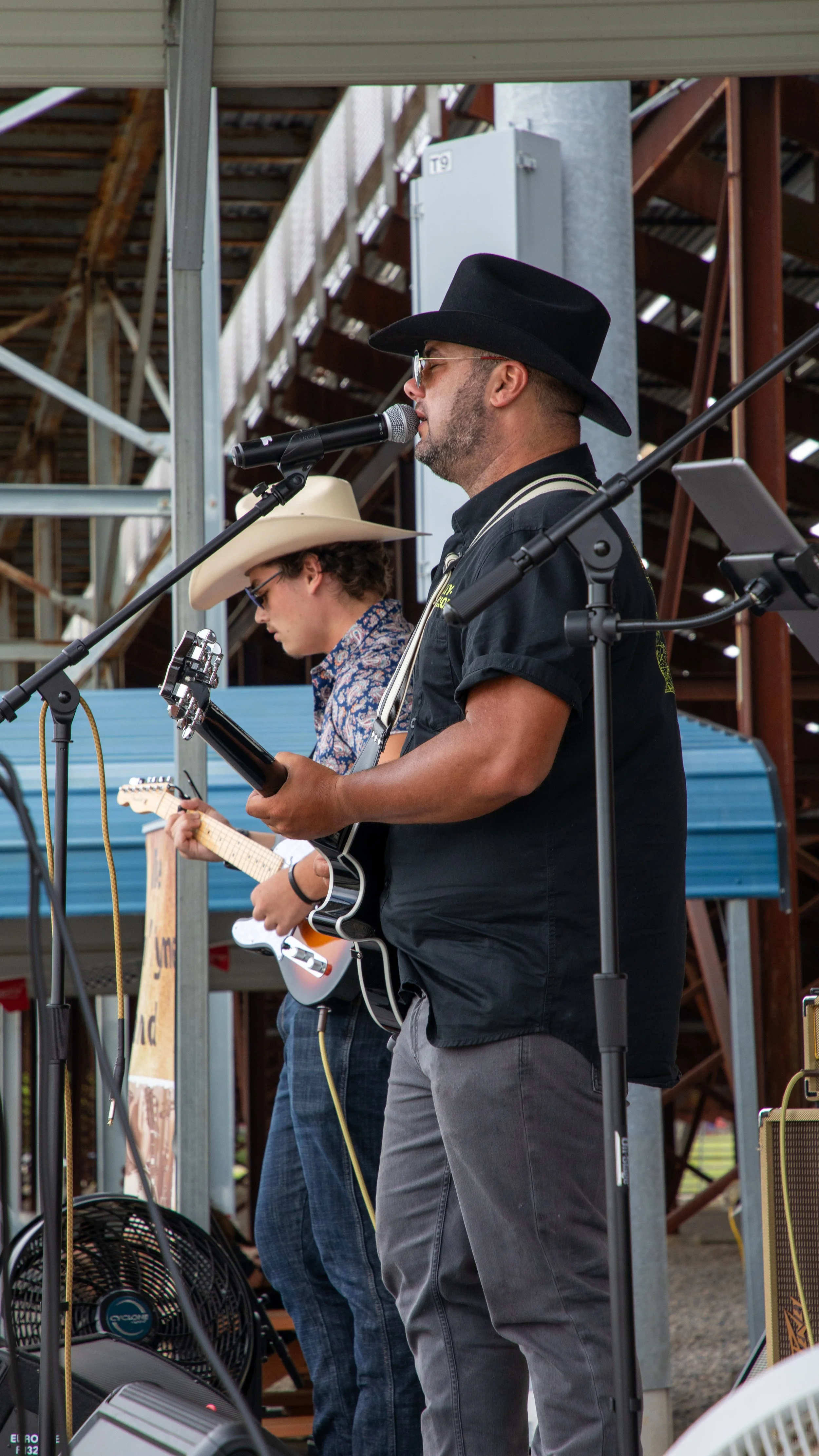 Concert Photography by Will Locke. Two musicians, one wearing a cowboy hat and the other a black hat, perform on stage with guitars and microphones at an outdoor event.