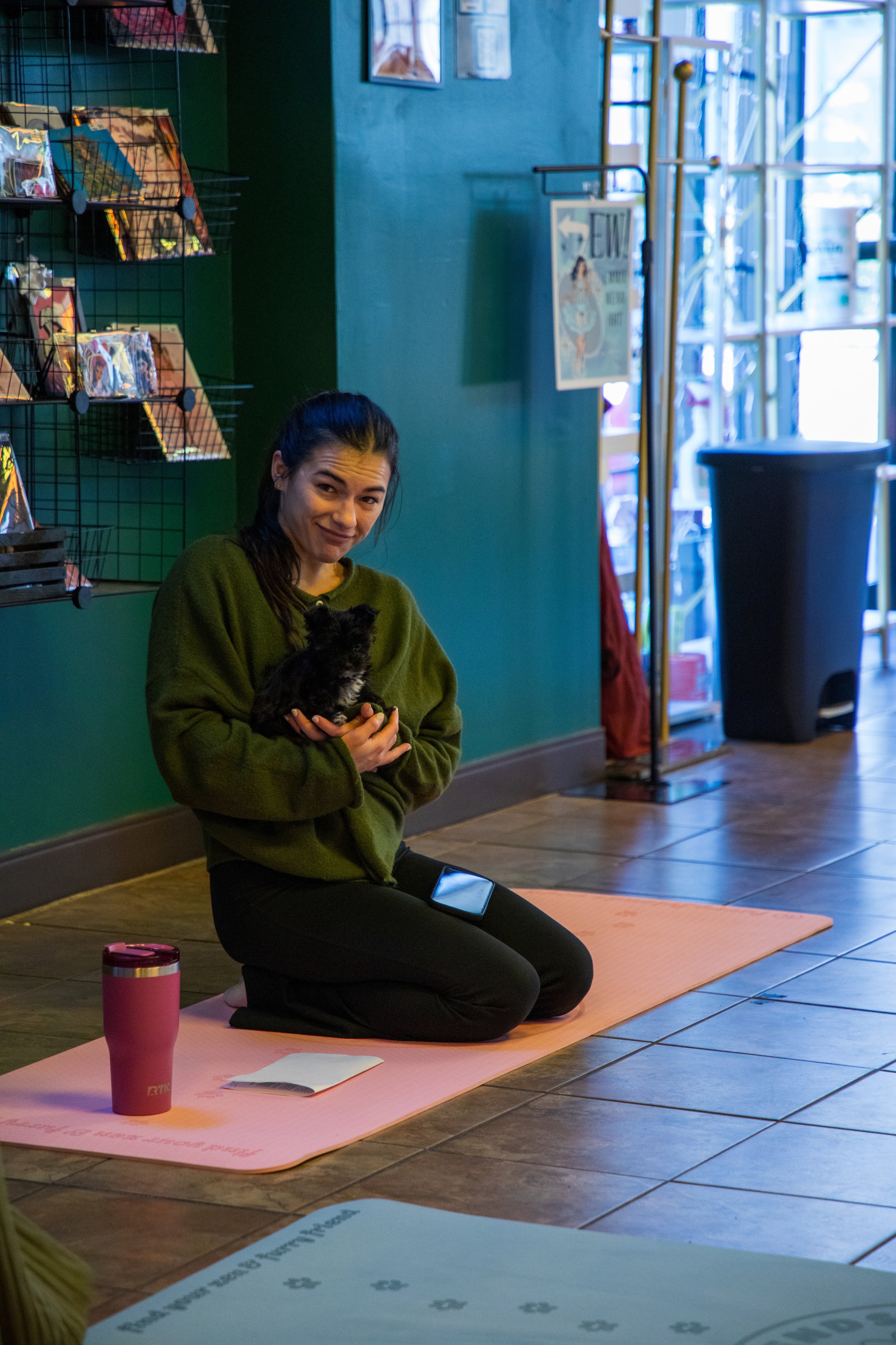 Marketing and Event Photography by Will Locke. A woman kneeling on a pink yoga mat indoors, holding a small black and white dog, with a green wall, display shelf with items, and a large window in the background.