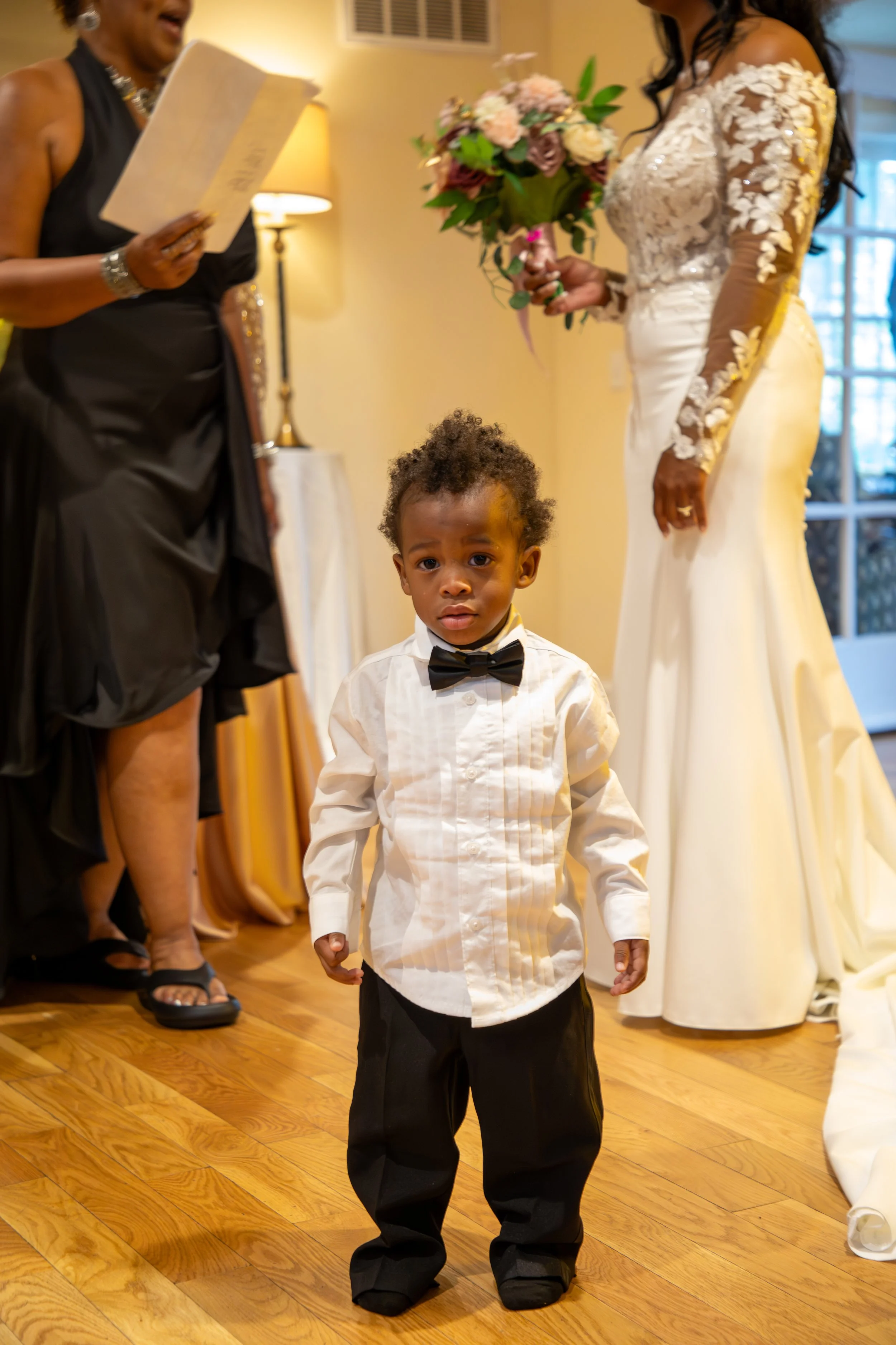 Photo & Video by Will Locke Wedding Photography at the Woman's Club of Portsmouth in VA. Young boy dressed in a white shirt with a black bow tie and black pants, with a bride holding a bouquet and a woman reading from a paper in the background.