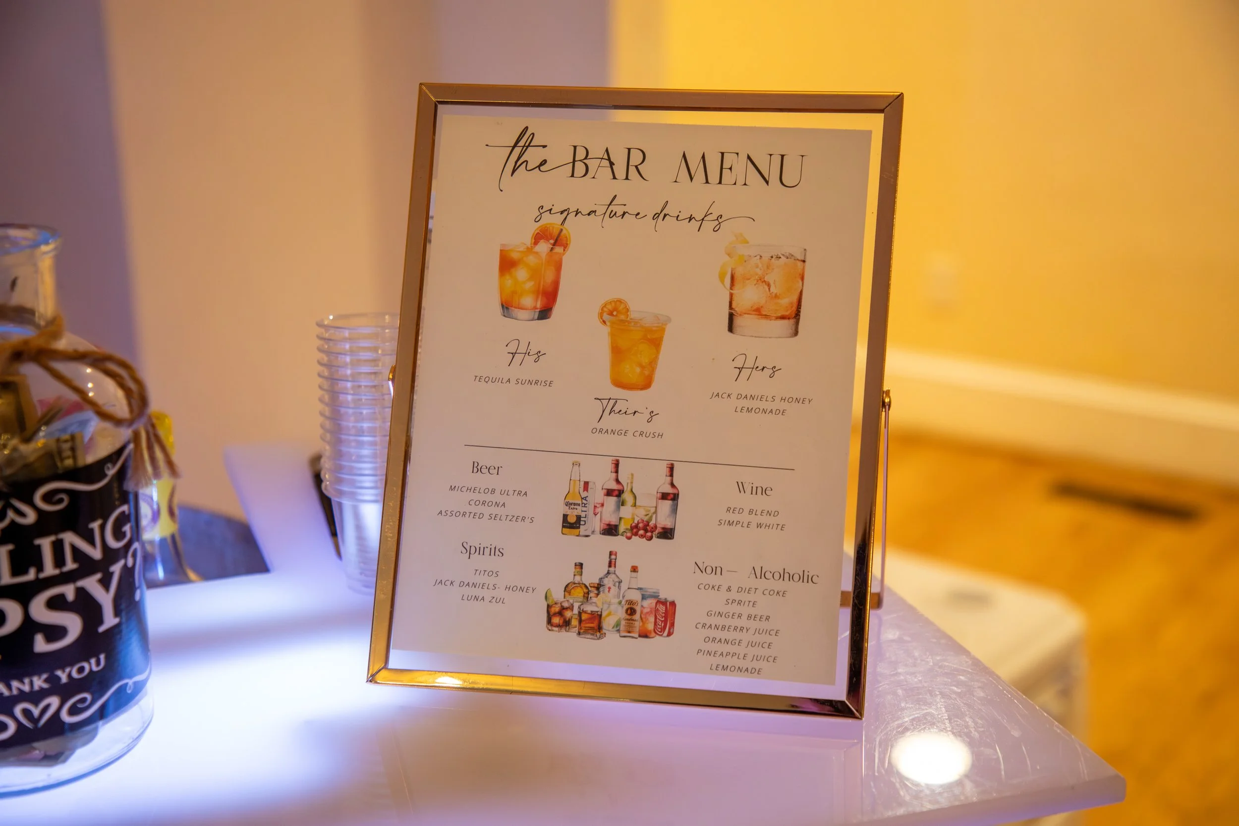 Wedding Photography by Will Locke. Framed bar menu featuring signature drinks, beers, spirits, wine, and non-alcoholic options on a white table with plastic cups and a drink jar labeled "Loving" with hearts.