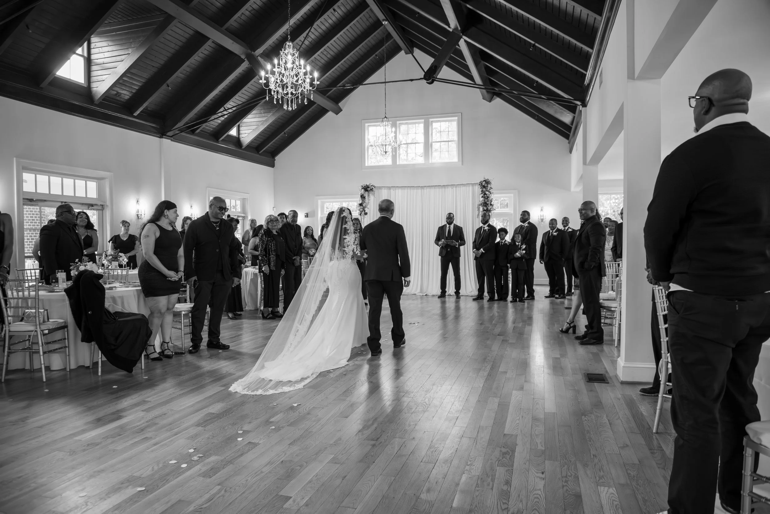 A bride and groom standing in the center of a wedding reception hall, surrounded by guests. The bride is wearing a long dress with a veil, and the groom is in a suit. The hall has high ceilings with a chandelier. Wedding Photography by Will Locke. 
