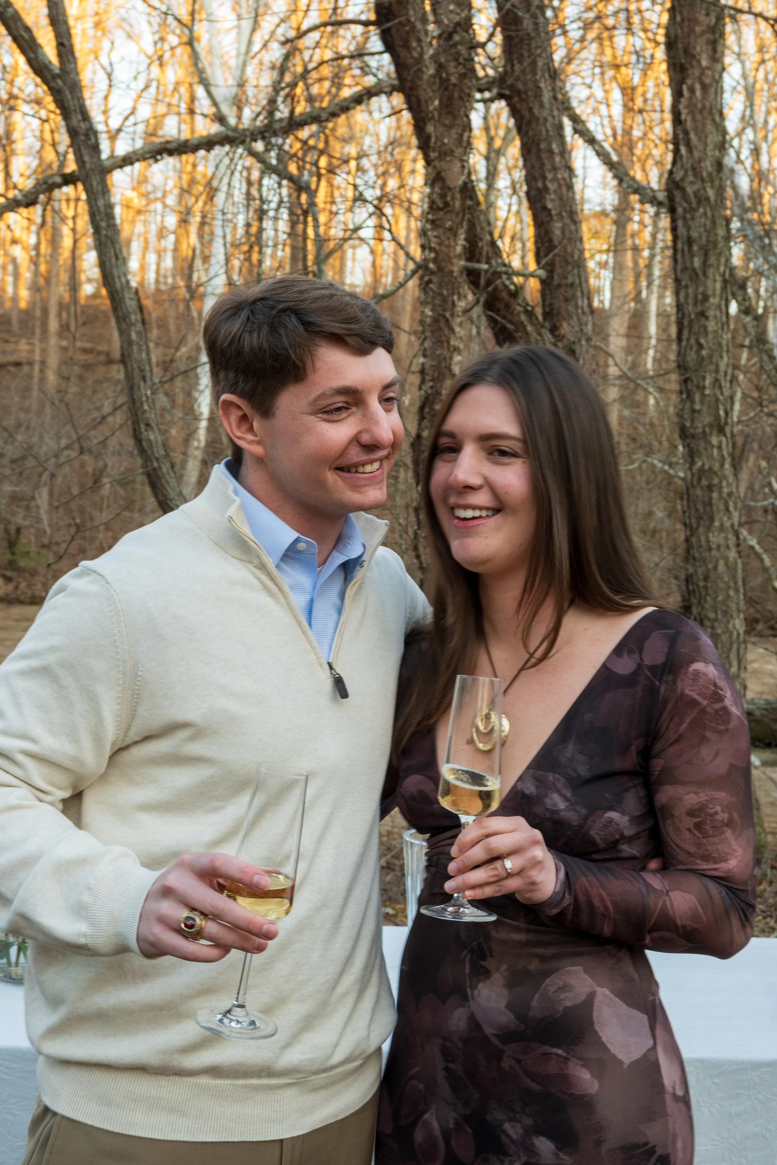 Engagement Photography by Will Locke near Richmond, VA in Montpelier. A man and woman smiling and hugging while holding champagne glasses outdoors during sunset, with a table of flowers nearby.