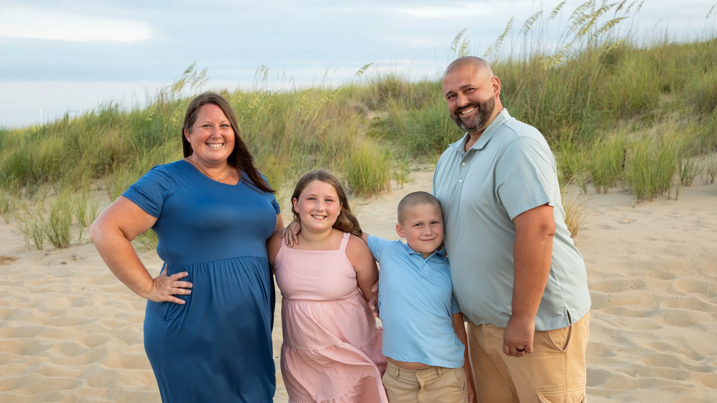 Photo & Video by Will Locke Family Photography in Virginia Beach, VA. Family of four posing on a sandy beach with dunes and grass in the background, smiling at the camera.