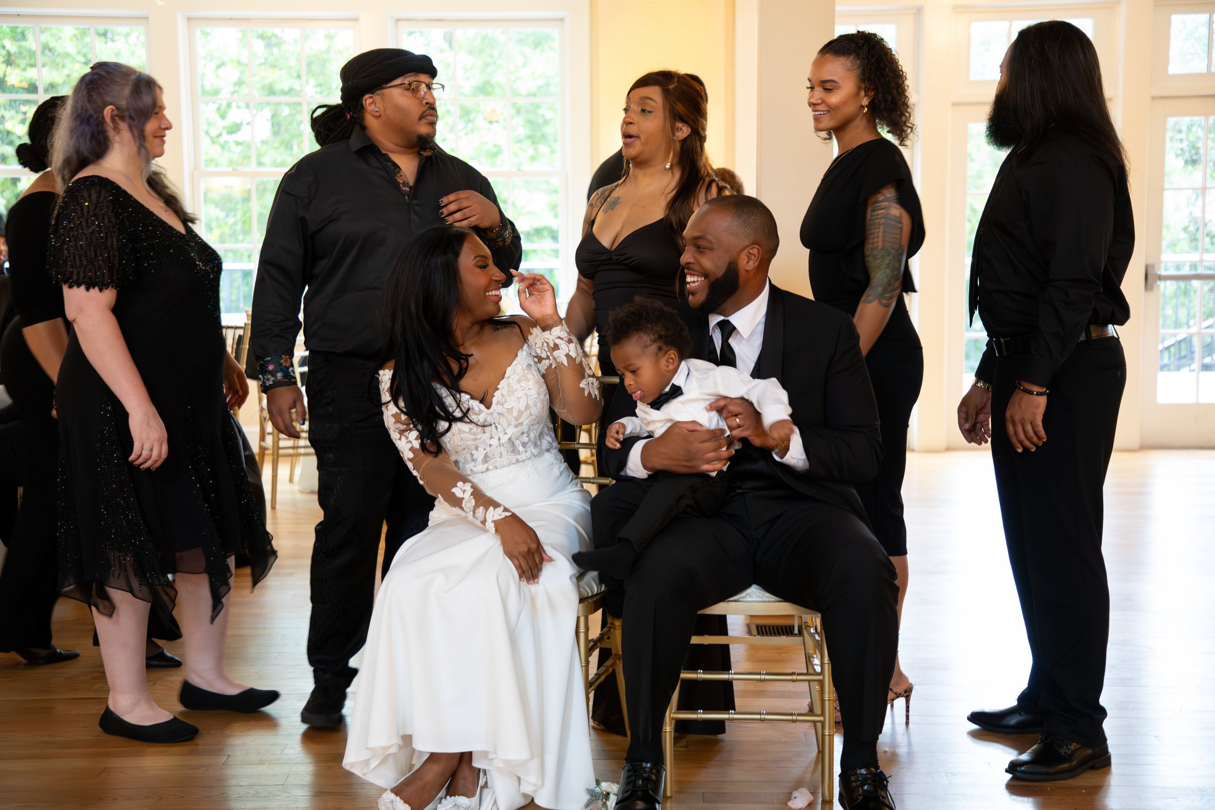A bride and groom are sitting together, celebrating their wedding with family and friends in a bright, indoor setting. The bride is wearing a white floral lace wedding gown. Photo & Video by Will Locke Wedding Photography. 