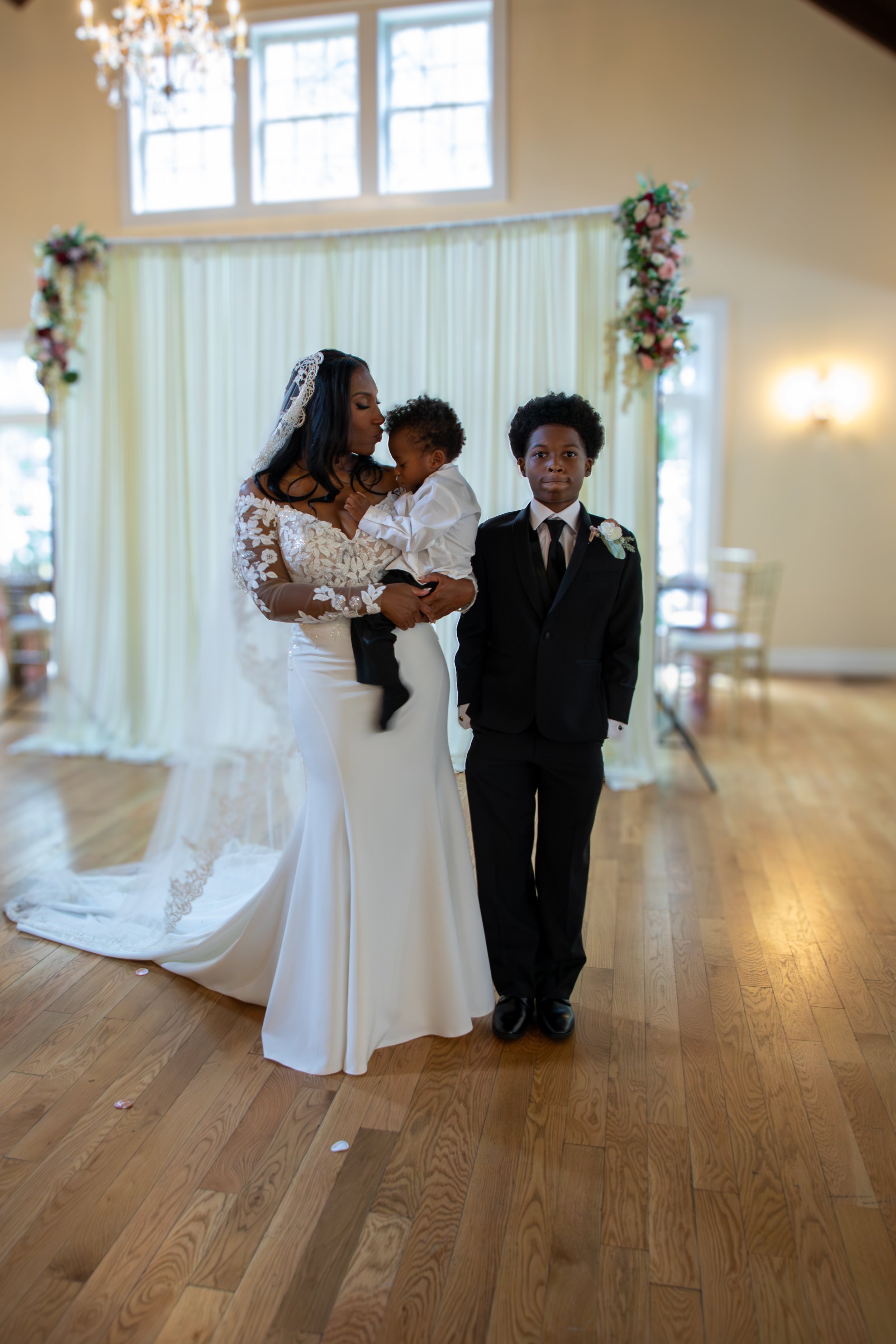 A bride holding a toddler in a white wedding dress with lace sleeves. Photo & Video by Will Locke Wedding Photography at the Woman's Club of Portsmouth in Virginia. 