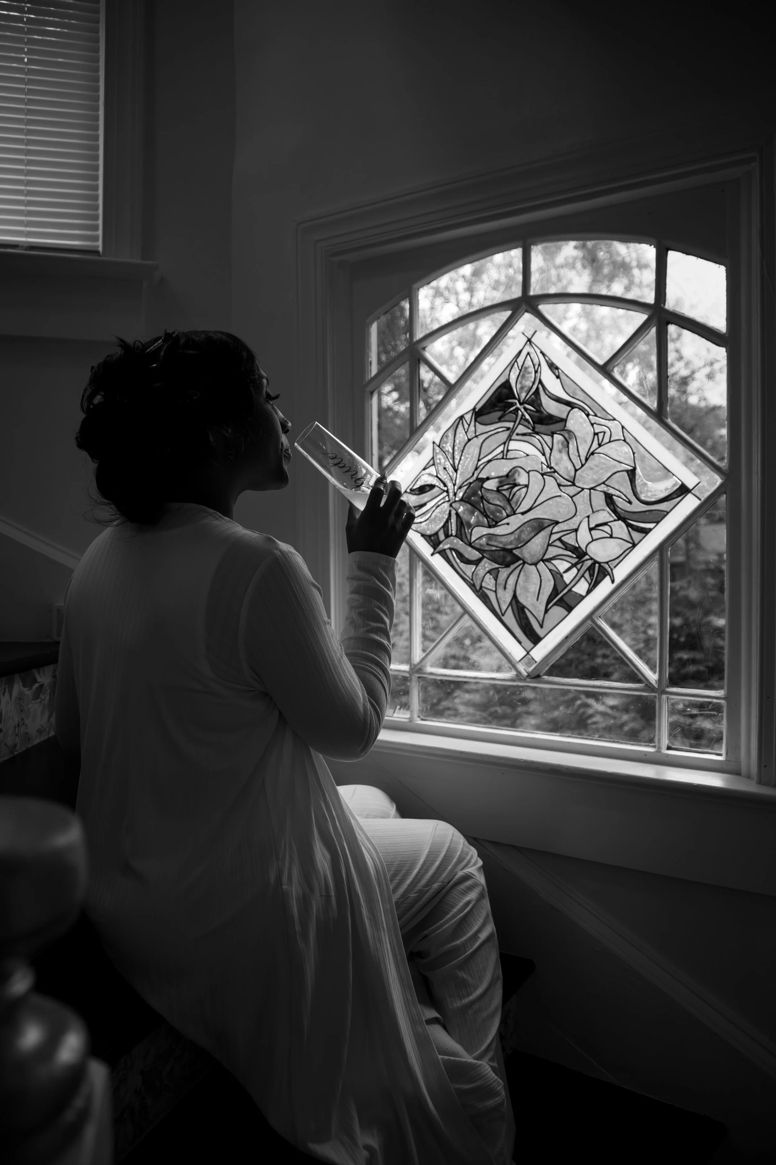 Photo & Video by Will Locke Wedding Photography at the Bridgerton House in Portsmouth, VA. A woman in light-colored clothing sitting on a window seat, drinking from a glass near a stained glass window with a floral design.