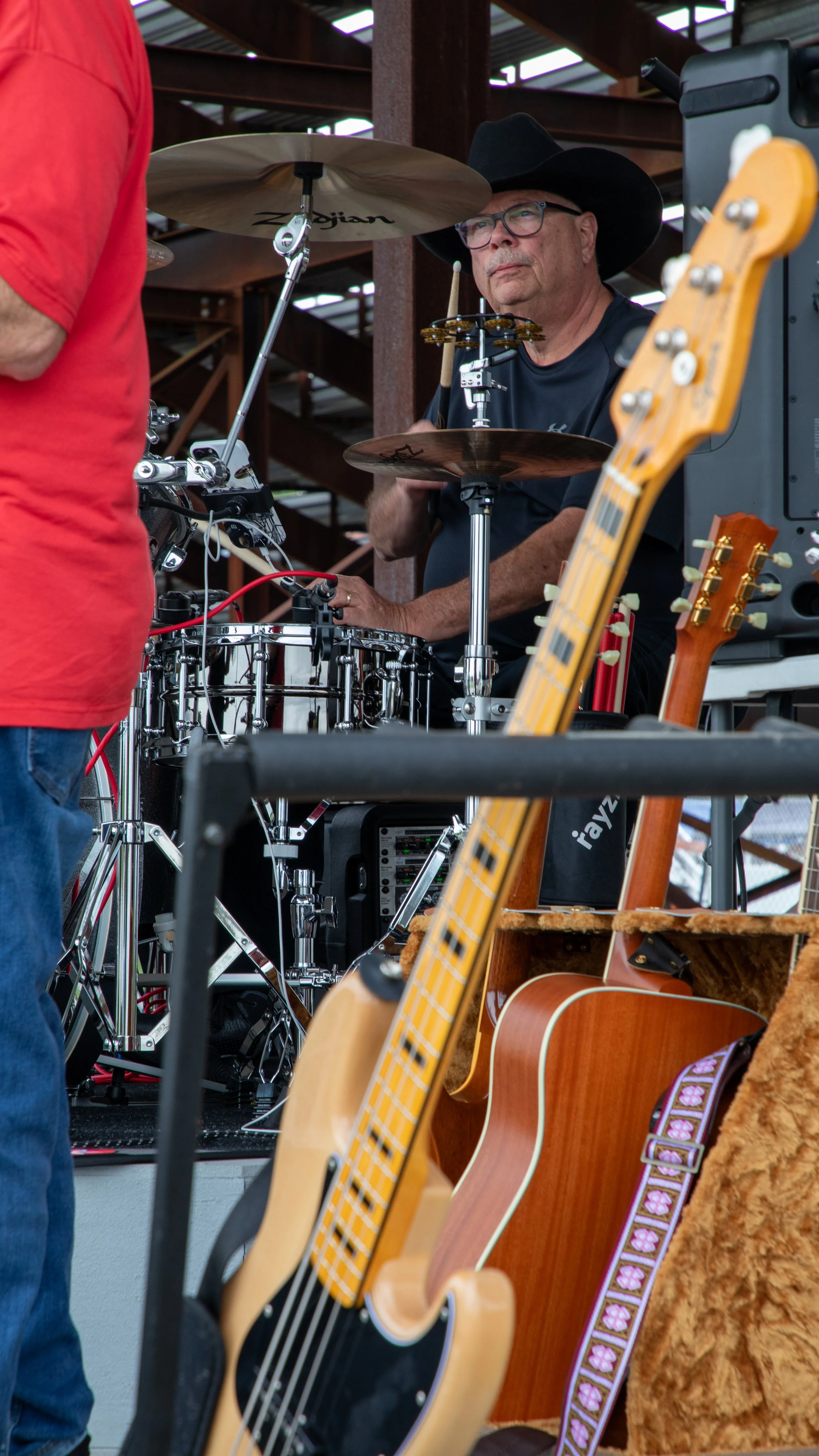 Concert Photography by Will Locke. A man wearing glasses and a cowboy hat playing a drum set behind guitar and other musical equipment at a performance