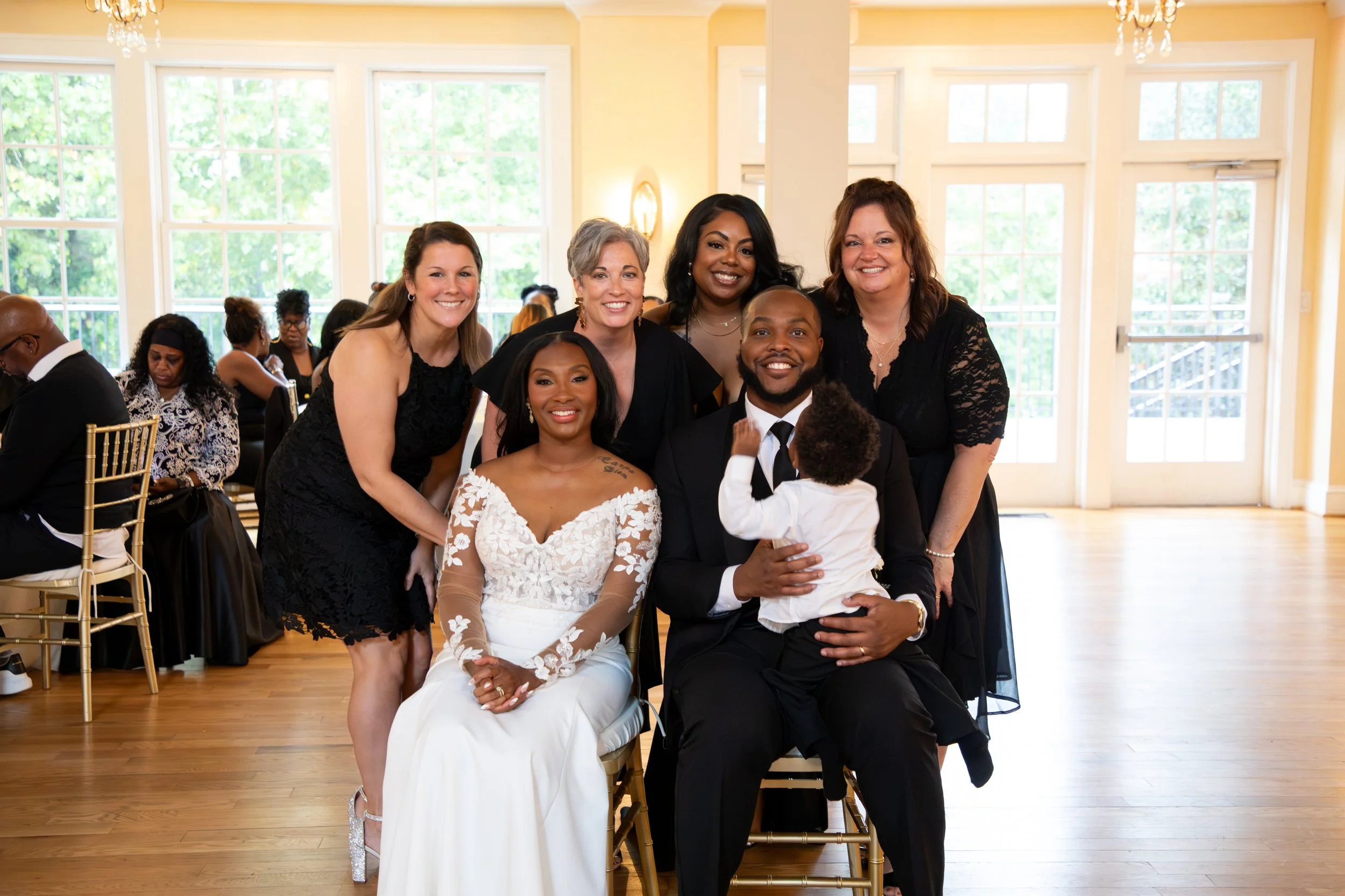 Photo & Video by Will Locke Wedding Photography at the Woman's Club of Portsmouth in Virginia. Group of six women and one man smiling at a wedding reception, with a woman in a white lace wedding gown seated.
