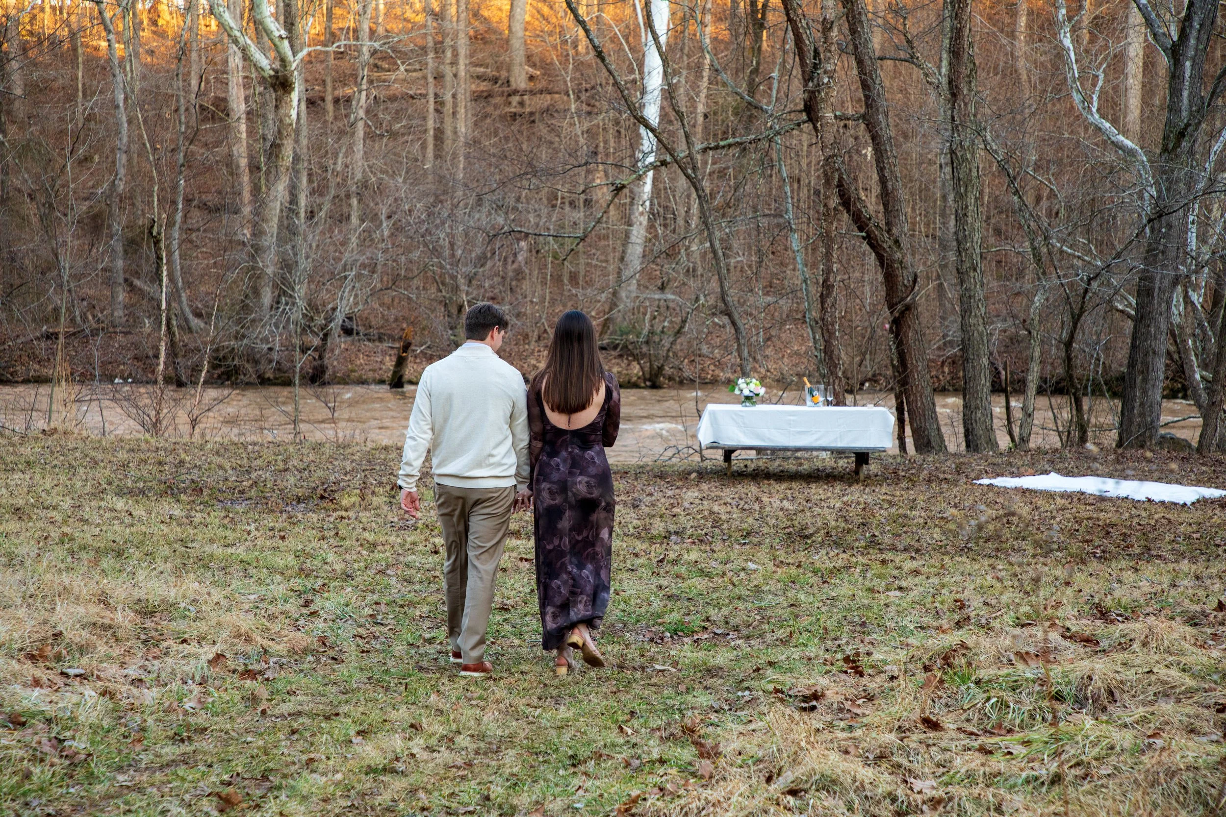 Engagement Photography by Will Locke near Richmond, VA in Montpelier. A couple walks to a white cloth on the ground near a wooded area with a stream on a fall day, with a table on the left side with white tablecloth.