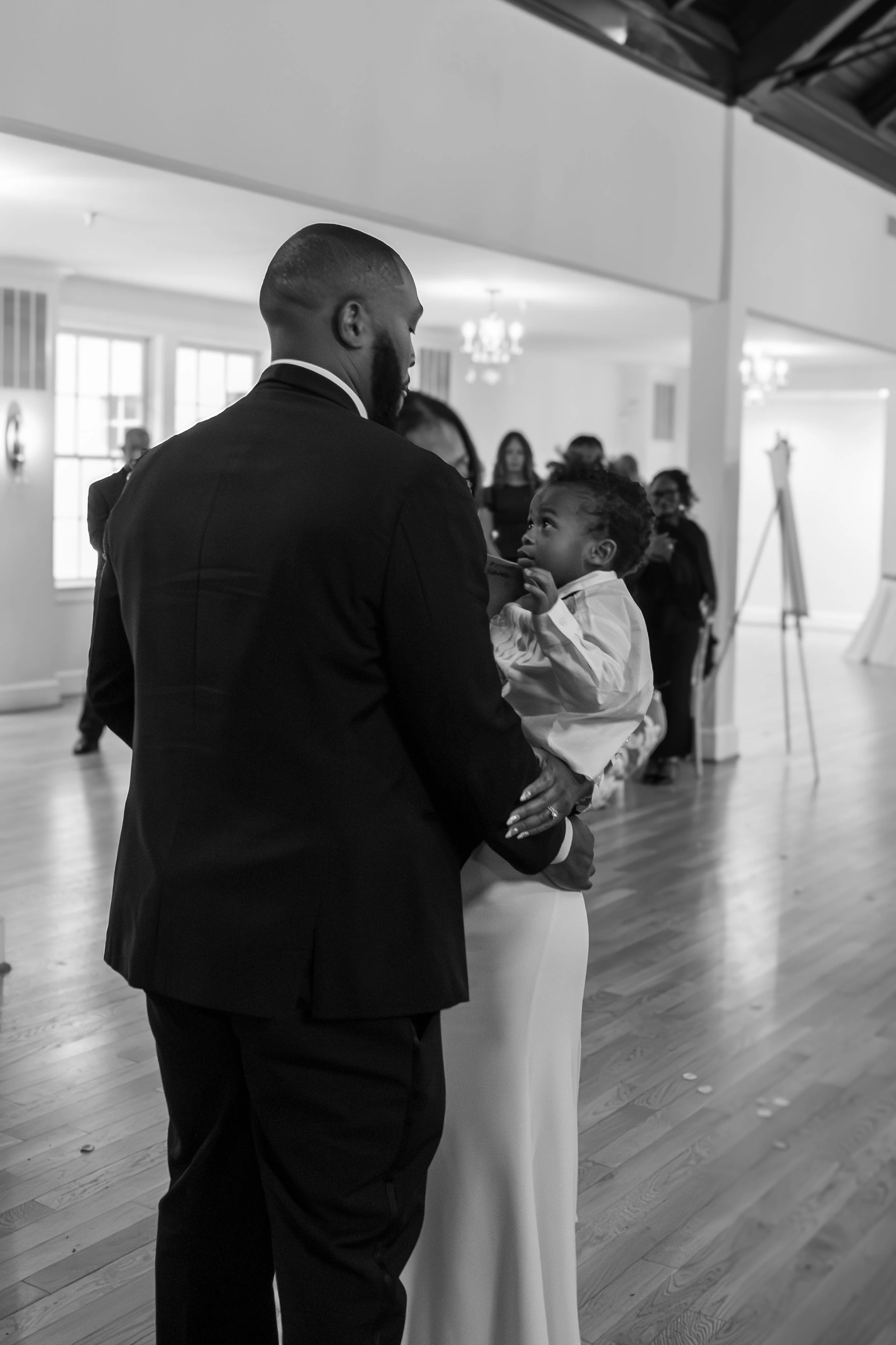 Photo & Video by Will Locke Wedding Photography at the Woman's Club of Portsmouth in Virginia. A black and white photo of a man in a suit holding a young girl, likely at a formal event or gathering, with people in the background.