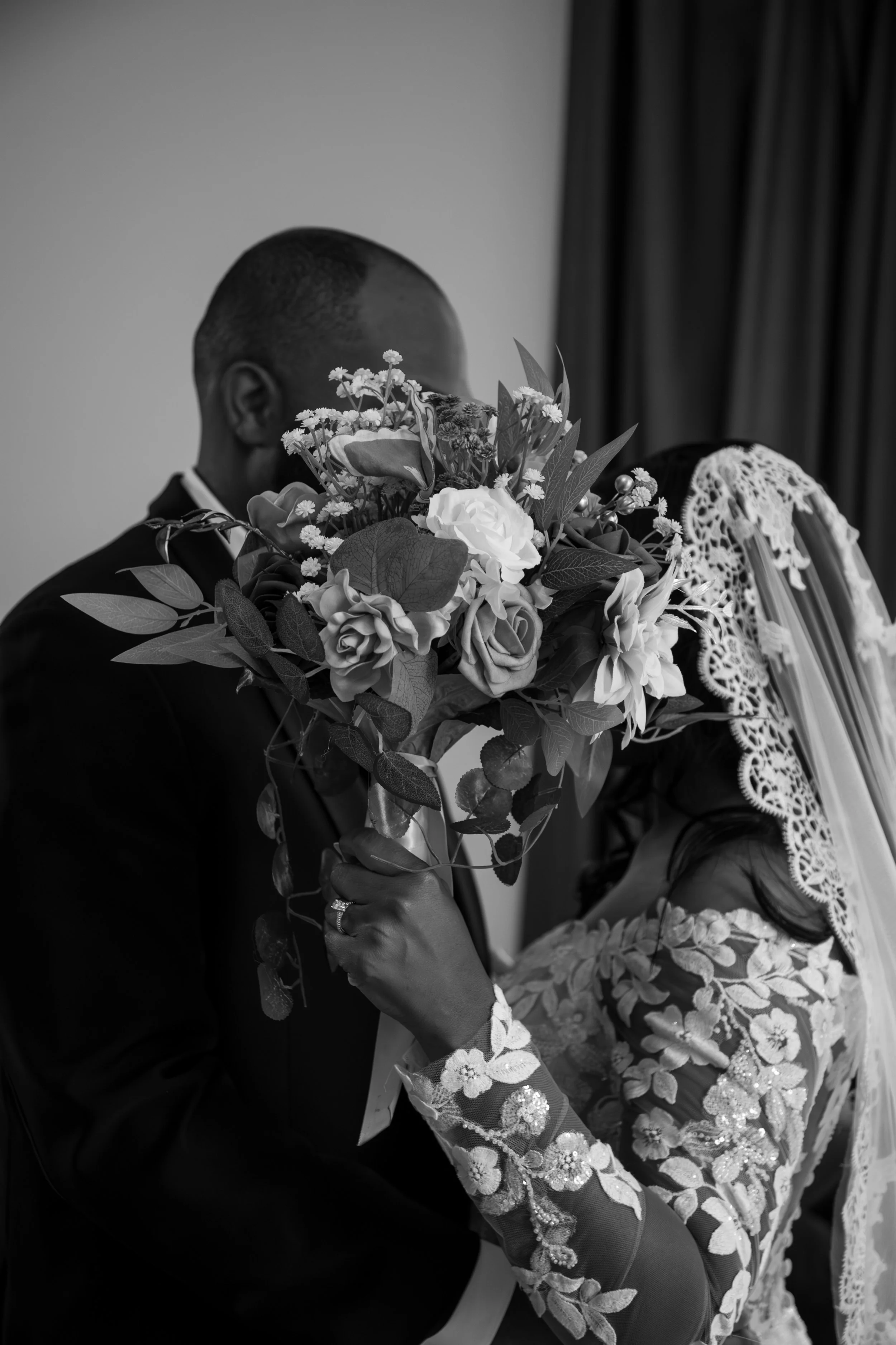 Wedding Photography by Will Locke. A black and white photograph of a bride and groom kissing, with the groom holding a bouquet of flowers in front of their faces. The bride is wearing a lace wedding dress with floral embroidery and a lace veil.