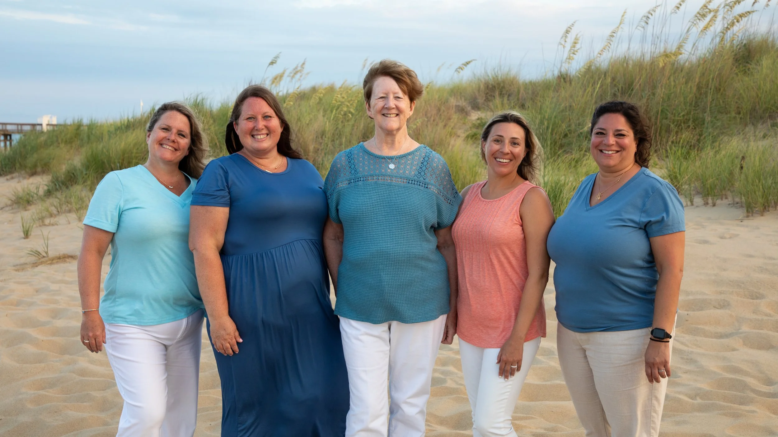 Photo & Video by Will Locke Family Photography in Virginia Beach, VA. Group of five women standing on a sandy beach with grass and dunes in the background, smiling at the camera.