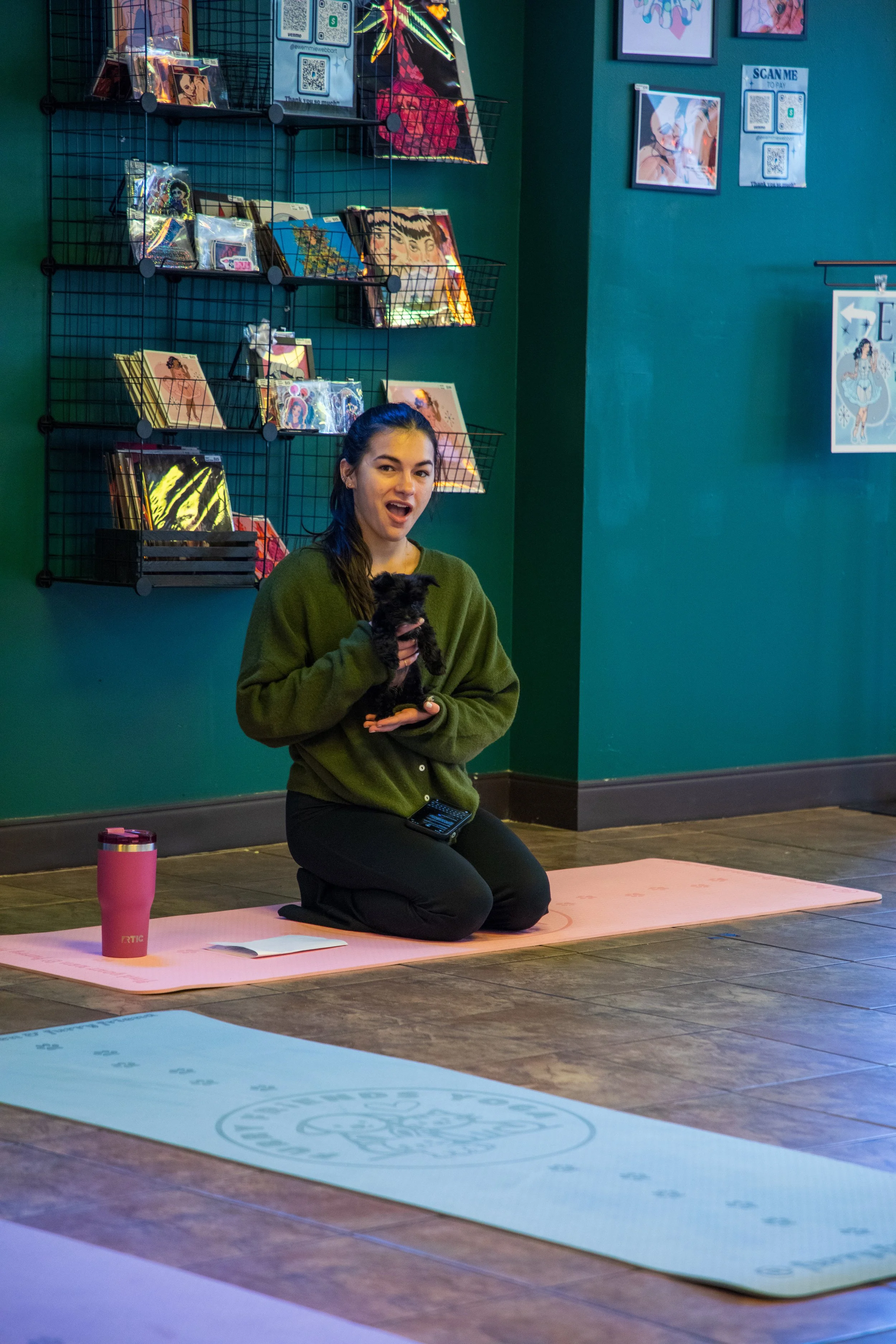 A young woman sitting on a pink yoga mat on the floor, holding a small black puppy, with a pink tumbler and a book on the mat. Marketing and Event Photography by Will Locke. 
