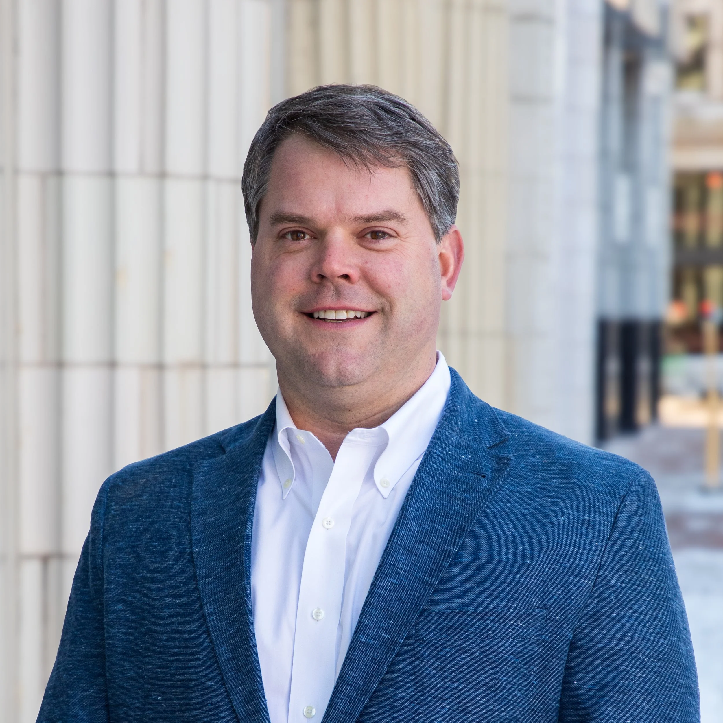 Photo & Video by Will Locke Commercial Headshot Photography for Canopy Development in Norfolk, Virginia. A man in a blue blazer and white shirt smiling outdoors in front of a building with large columns and glass windows.