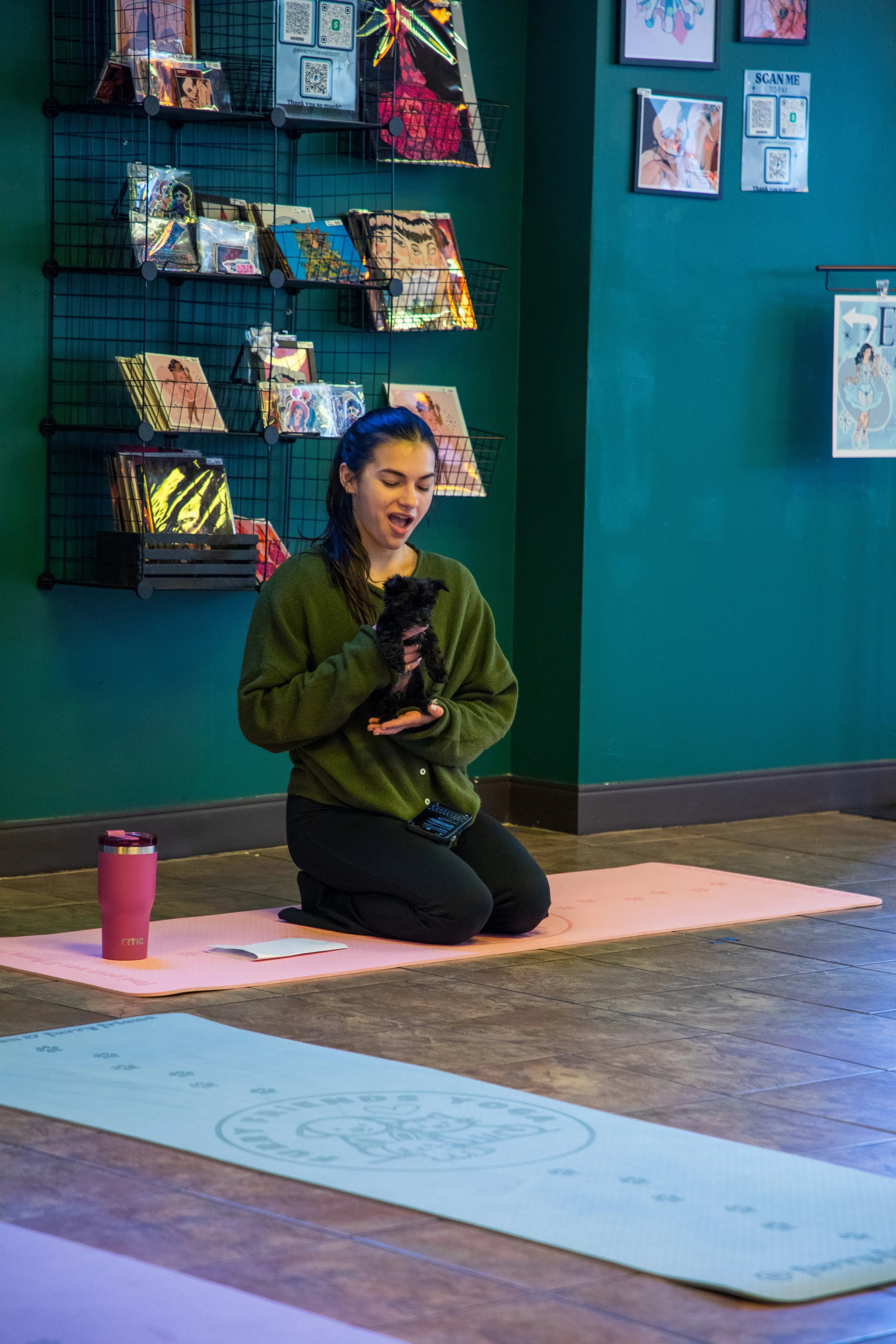 A woman kneeling on a pink yoga mat, holding a small black dog, with a pink tumbler and a notebook on the mat. Marketing and Event Photography by Will Locke. 