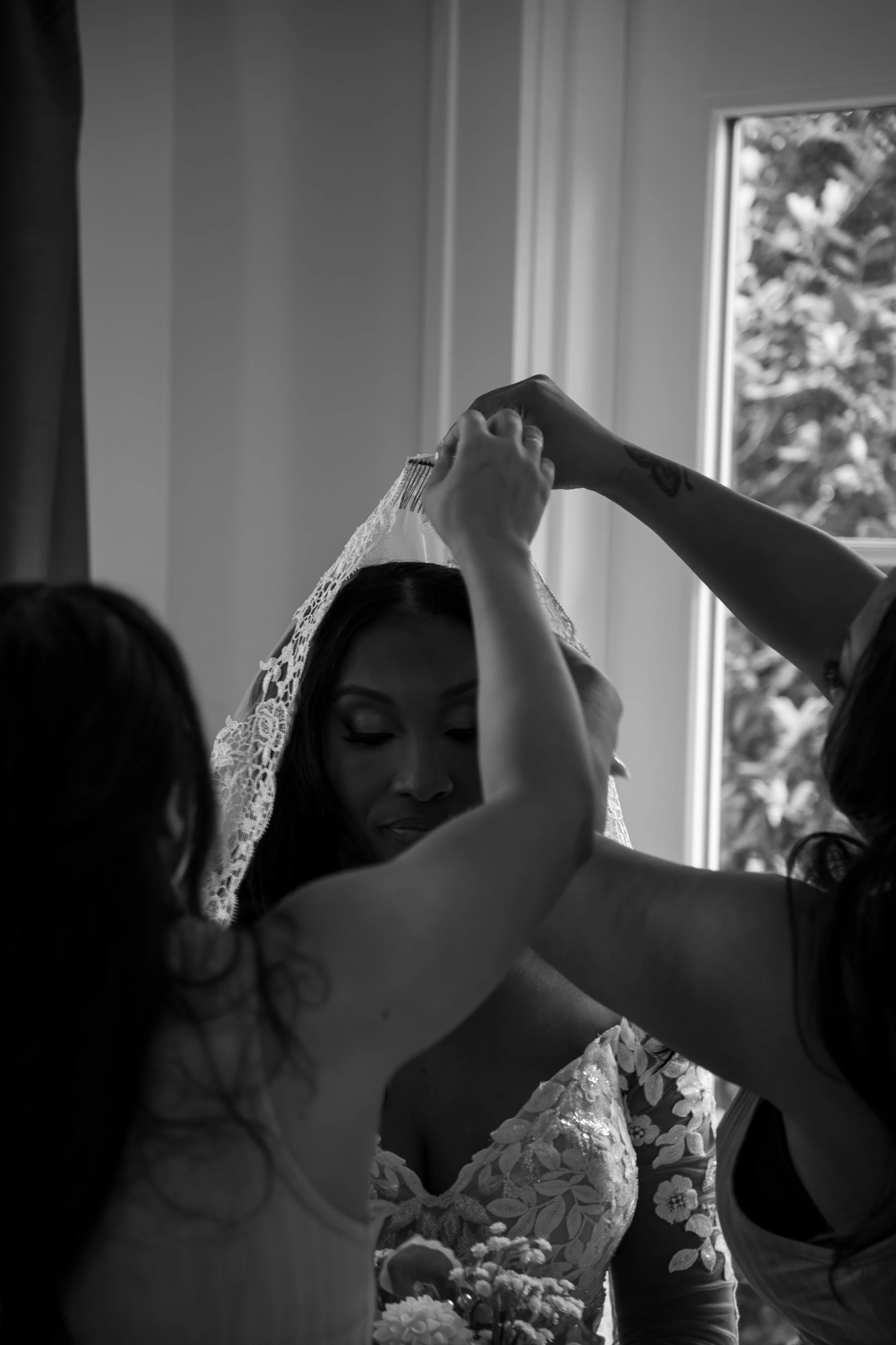 Wedding Photography by Will Locke. A bride is being prepared by her bridesmaids as they place her lace veil on her head, near a window with visible trees outside in the background.