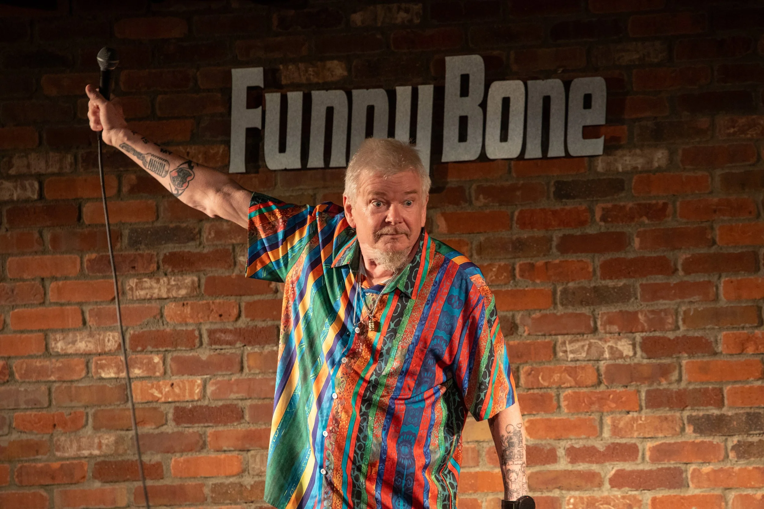 Photo & Video by Will Locke Comedy Photography. A comedian on stage at the Funny Bone comedy club, holding a microphone in his right hand, wearing a colorful, patterned shirt, with a brick wall and the 'Funny Bone' sign behind him.