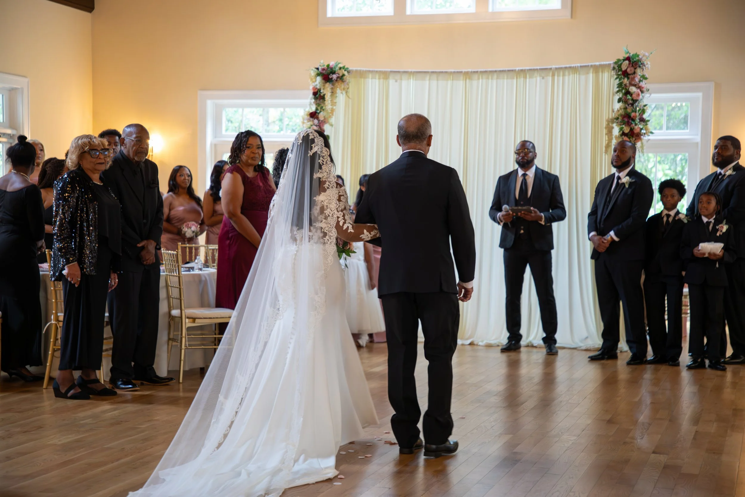 Wedding Photography by Will Locke. A wedding ceremony with a bride in a white gown and veil walking down the aisle, escorted by a man in a black suit, in a decorated hall with a cream-colored curtain and floral arrangements.