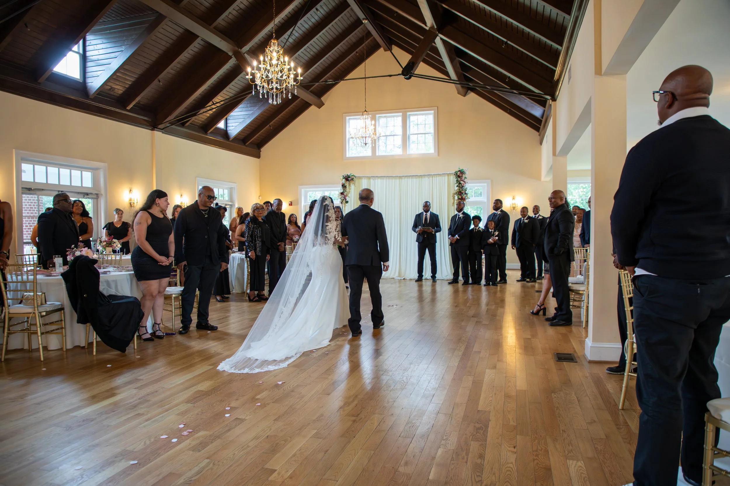 A wedding ceremony in a spacious hall with a high wooden ceiling, chandeliers, and large windows. Photo & Video by Will Locke Wedding Photography. 