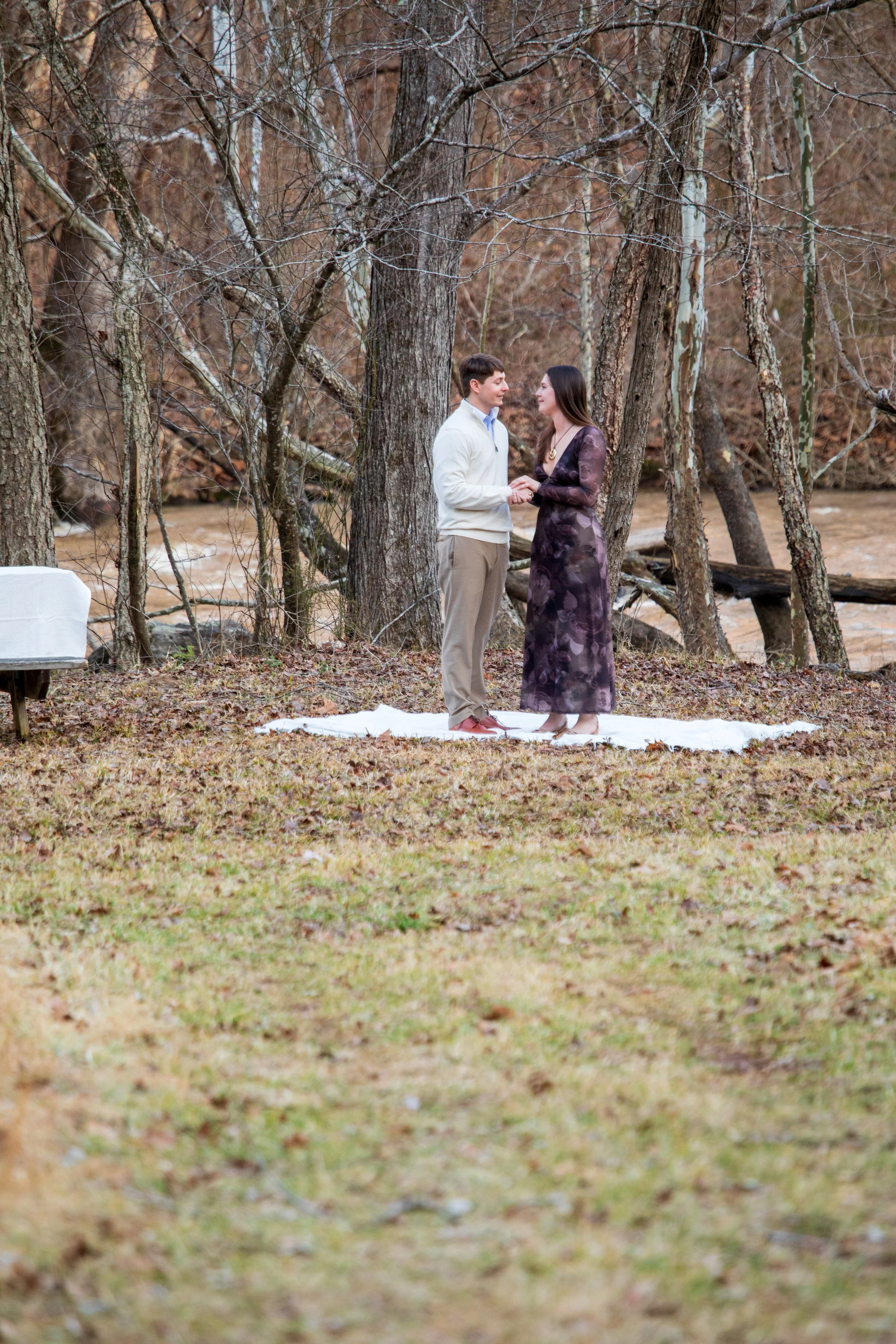 Engagement Photography by Will Locke near Richmond, VA in Montpelier. A couple standing on a white cloth outdoors, holding hands, with trees and a river in the background.