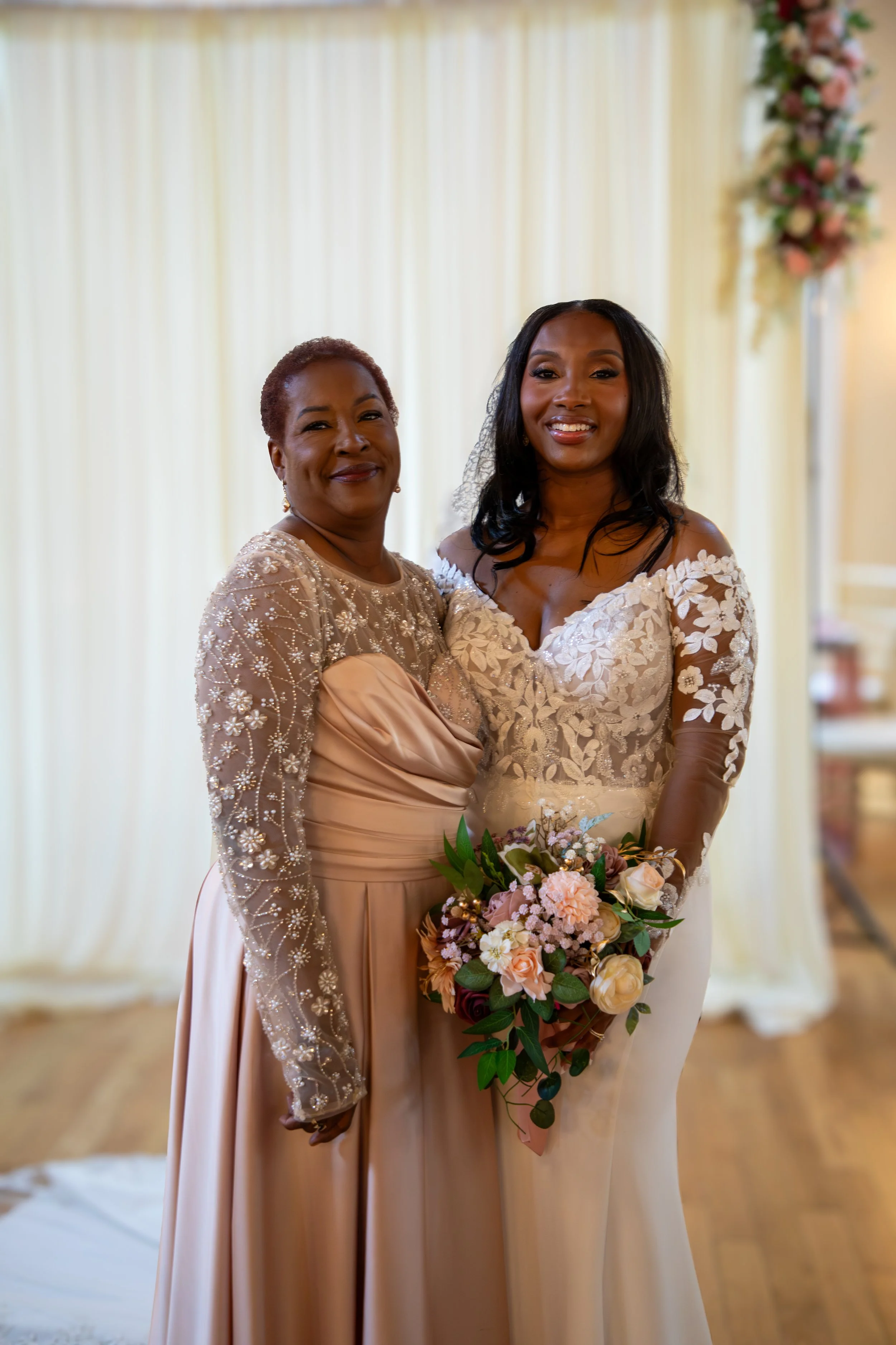 Photo & Video by Will Locke Wedding Photography at the Woman's Club of Portsmouth in VA. Two women in elegant dresses smiling during a wedding ceremony, with one holding a bouquet of flowers.